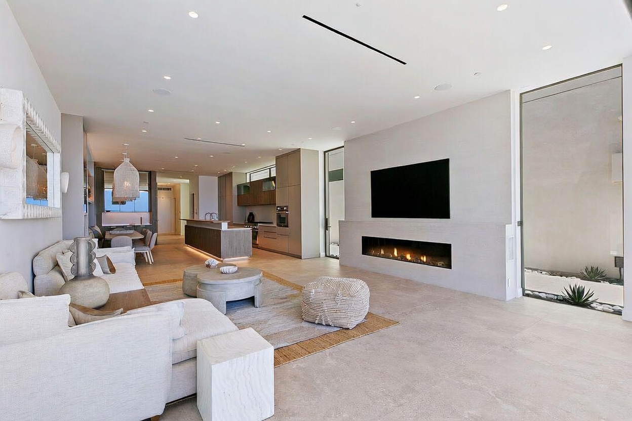 This open-concept living area features a neutral color palette with light-toned tile flooring and a textured area rug. A large television is mounted above a linear fireplace set into a prominent wall, while a comfortable sofa and circular coffee tables create a welcoming seating arrangement. The space flows seamlessly into a modern kitchen in the background, with large windows providing natural light and a view of an outdoor area.