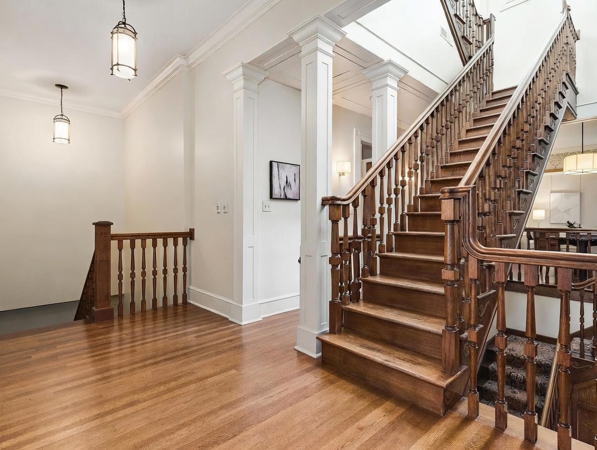 This elegant foyer features a grand wooden staircase with intricate balusters and a polished handrail, serving as the central architectural focal point. The space is finished with light-toned walls, white columns, and warm-toned wood flooring that extends throughout the entryway. A baby gate is installed at the base of the stairs, and a pendant light fixture hangs from the ceiling, creating a welcoming and traditional atmosphere.