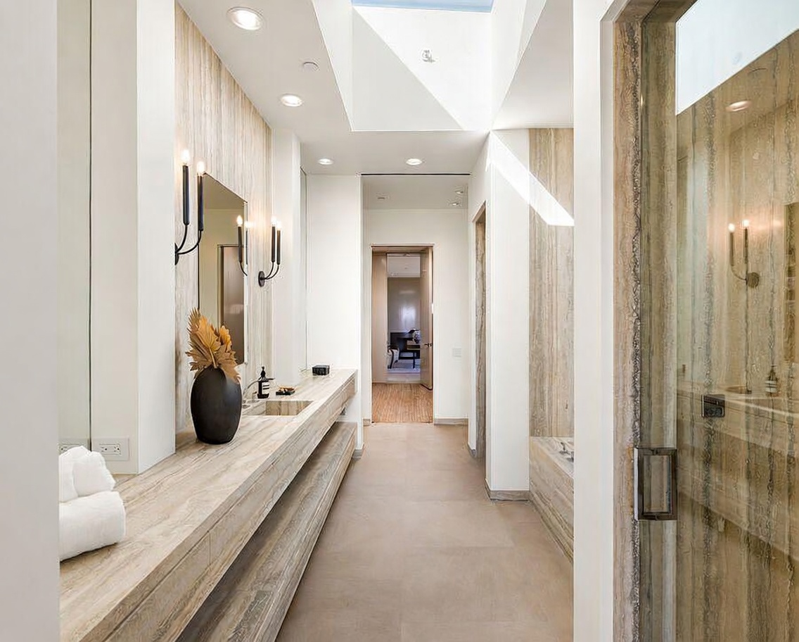 This luxurious primary bathroom features a long, floating vanity crafted from elegant, light-toned travertine stone, complemented by matching wall accents and a glass-enclosed shower. A skylight above the hallway floods the space with natural light, highlighting the clean, modern lines and sophisticated neutral color palette. The perspective looks down the hallway, showcasing the seamless transition between the vanity area and the adjacent living space.