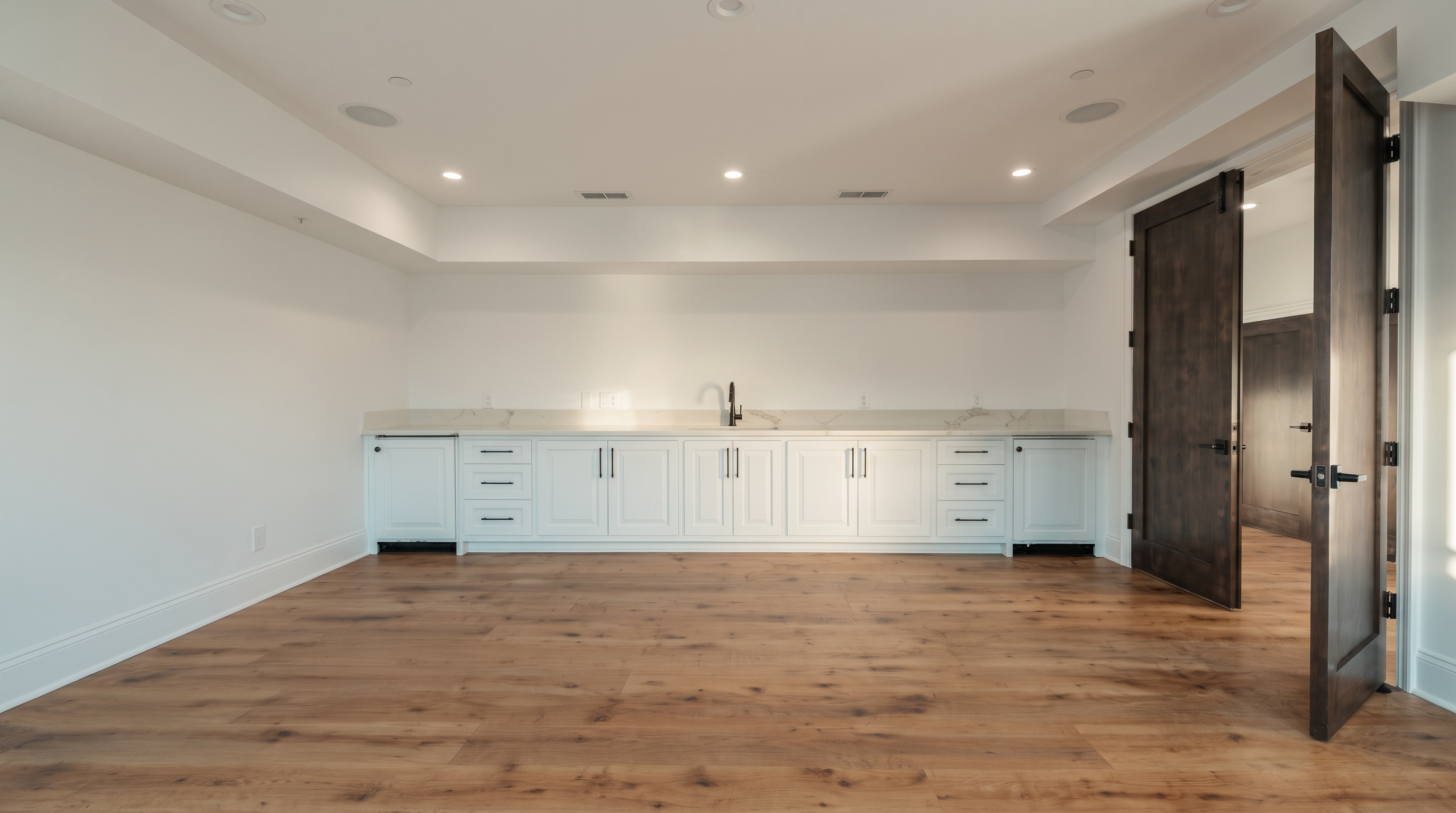 This spacious, minimalist room features light wood flooring and crisp white walls, creating a bright and versatile living area. A long, built-in cabinetry unit with a central sink and faucet spans the back wall, offering ample storage and utility. To the right, a set of dark, double-hinged doors adds a sophisticated contrast to the otherwise neutral palette of the room.