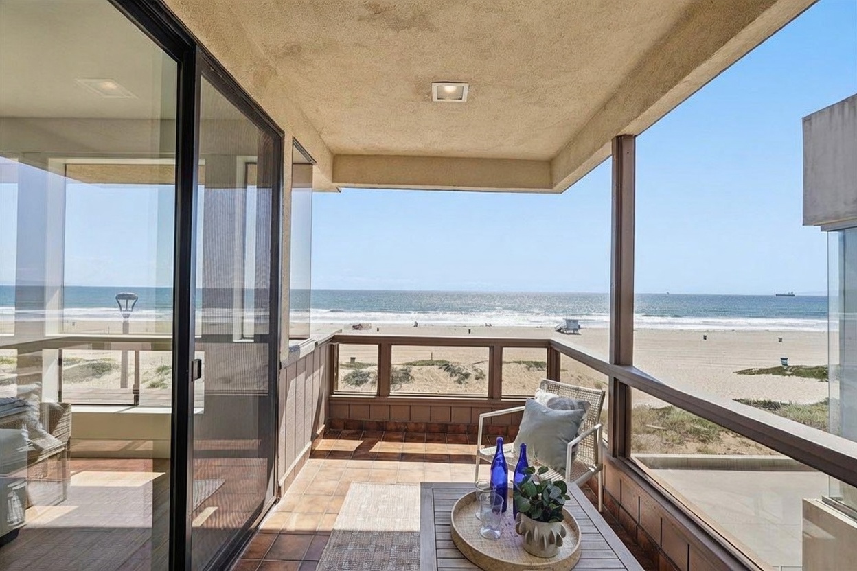 This balcony offers a direct, unobstructed view of the ocean and sandy beach, creating a serene and luxurious atmosphere. The space is furnished with a small wooden table holding a decorative tray and blue glass bottles, alongside a comfortable chair with a plush pillow. Large sliding glass doors provide seamless indoor-outdoor flow, while the tiled floor and wooden railing define the private outdoor living area.
