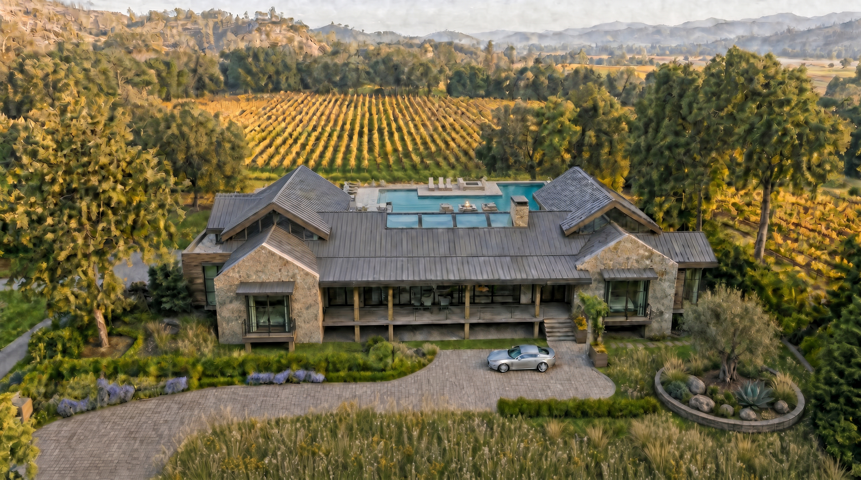 This stunning luxury estate is captured from an elevated perspective, showcasing a sprawling stone-clad residence nestled within a vast vineyard. The house features a metal roof, an integrated infinity-edge swimming pool with an outdoor lounge area, and a private driveway leading to the grand entrance. The surrounding landscape, abundant with mature trees and formal garden accents, creates a serene and sophisticated atmosphere.