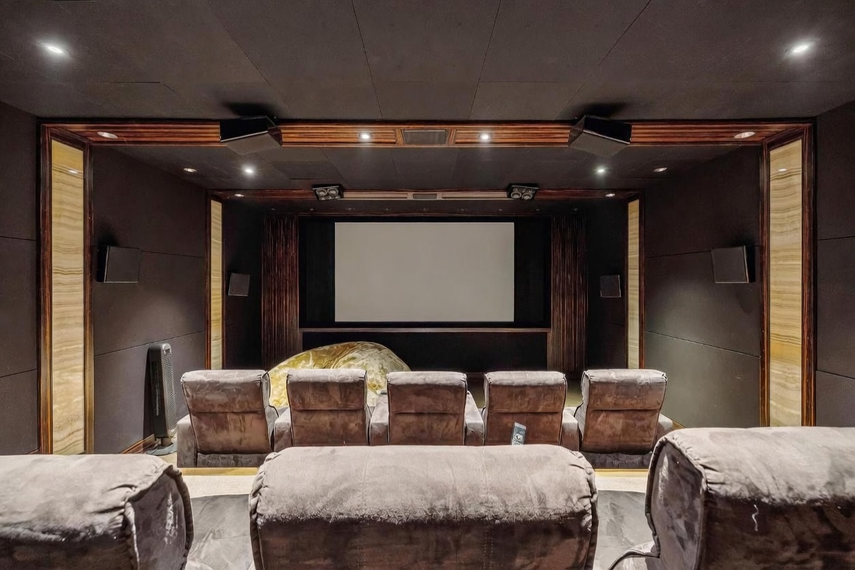 This dedicated home theater room features tiered rows of plush, dark-toned reclining seating facing a large projection screen. The walls are finished with dark acoustic paneling and accented by vertical light-wood panels, while recessed ceiling lights and wall-mounted speakers create an immersive cinematic environment. The perspective is from the back of the room, looking forward toward the screen, emphasizing the professional layout and comfortable viewing experience.
