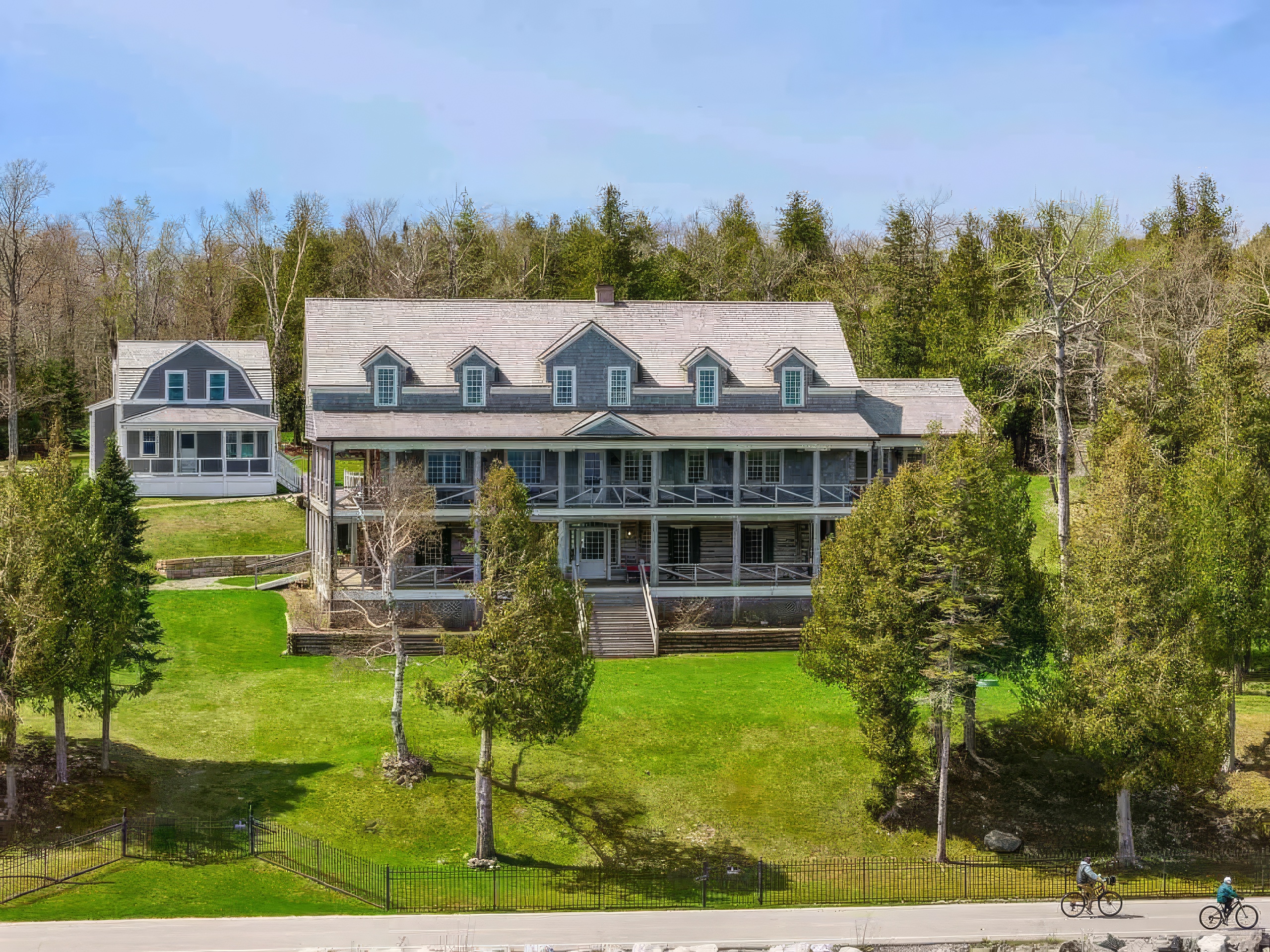 This elevated perspective captures a large, multi-story shingle-style home featuring expansive covered porches on both the main and upper levels. The property is set against a backdrop of dense, mature trees and includes a smaller, matching secondary structure to the left. A manicured lawn slopes gently toward a black metal fence, with a paved path in the foreground where two cyclists are visible.