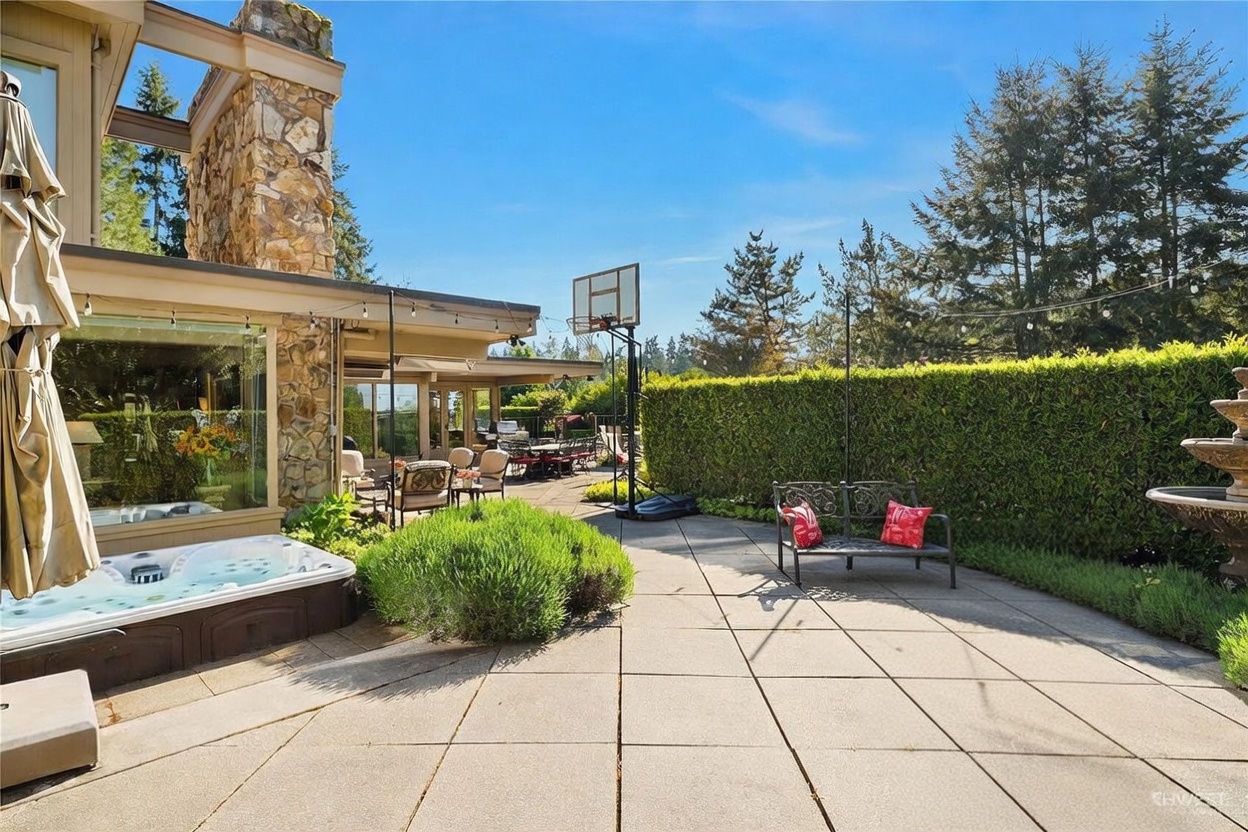 This expansive stone-paved patio offers a serene outdoor living space, featuring a built-in hot tub and a comfortable seating area with wrought-iron furniture. A tall stone-clad chimney breast anchors the side of the house, while a manicured hedge provides privacy and a lush backdrop. The scene is captured from a ground-level perspective, highlighting the seamless transition between the home's interior and the sunny, well-maintained backyard.