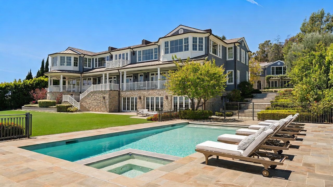 This expansive rear view of a luxury estate showcases a multi-level residence with a classic architectural style, featuring a stone-clad lower level and dark-toned siding above. A pristine swimming pool with an integrated spa takes center stage in the foreground, surrounded by a stone patio lined with lounge chairs. The property includes a manicured lawn, mature trees, and a white-railed balcony that overlooks the outdoor living space.