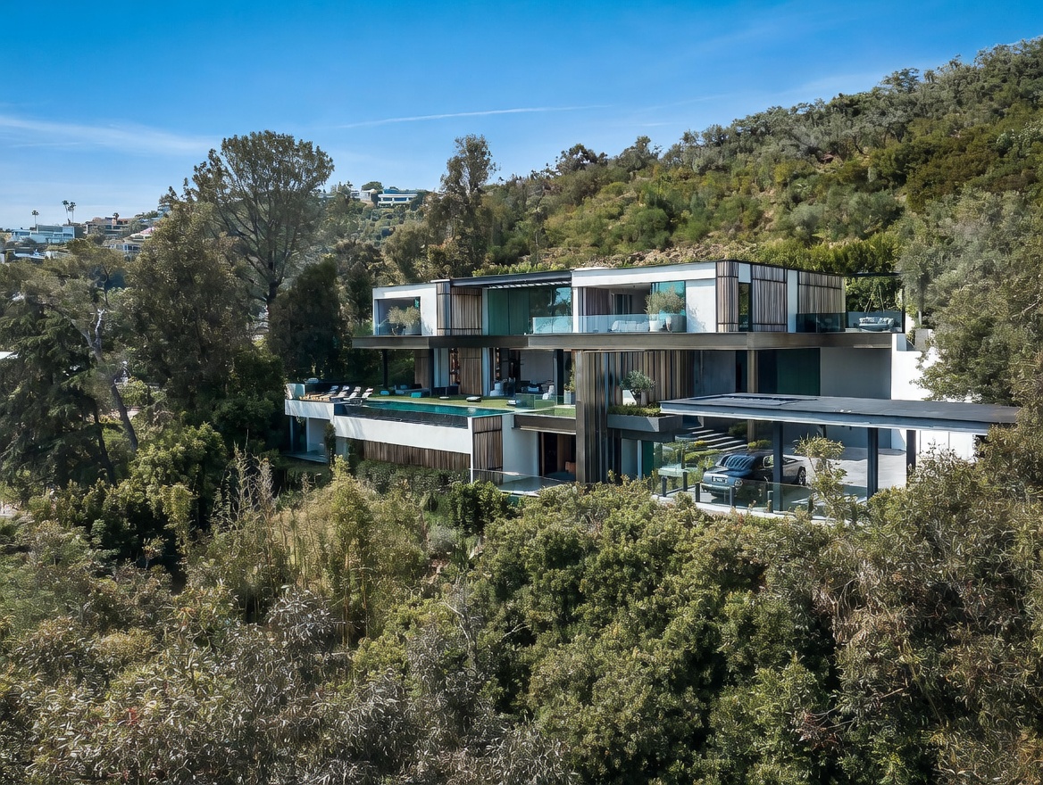 This stunning contemporary architectural masterpiece is captured from an elevated aerial perspective, showcasing its multi-level design integrated into a lush, hillside landscape. The home features clean lines, extensive glass walls, a sleek infinity-edge pool, and a covered carport, all set against a backdrop of dense greenery and a clear blue sky. The overall impression is one of luxury, privacy, and sophisticated modern living.