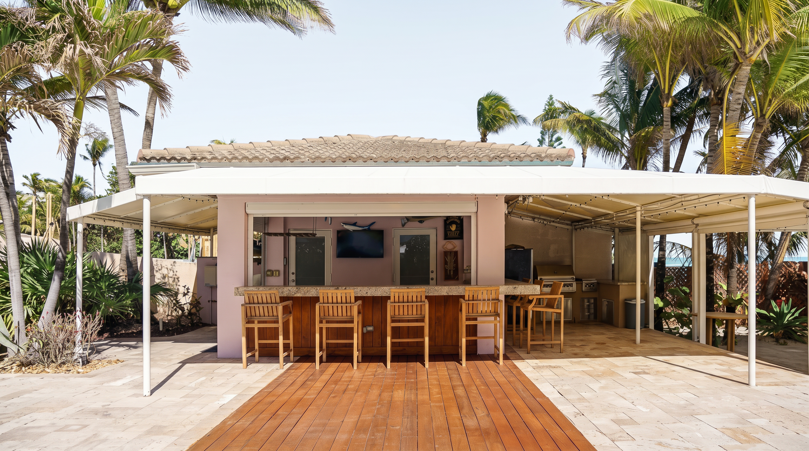 This outdoor living space features a covered bar area with a stone-topped counter and five wooden bar stools, set against a backdrop of lush palm trees. The area includes a television, a grill station, and a wooden deck transition leading to a paved patio, creating an ideal setting for tropical entertaining. The open-air design provides a relaxed, resort-style atmosphere perfect for enjoying the coastal environment.