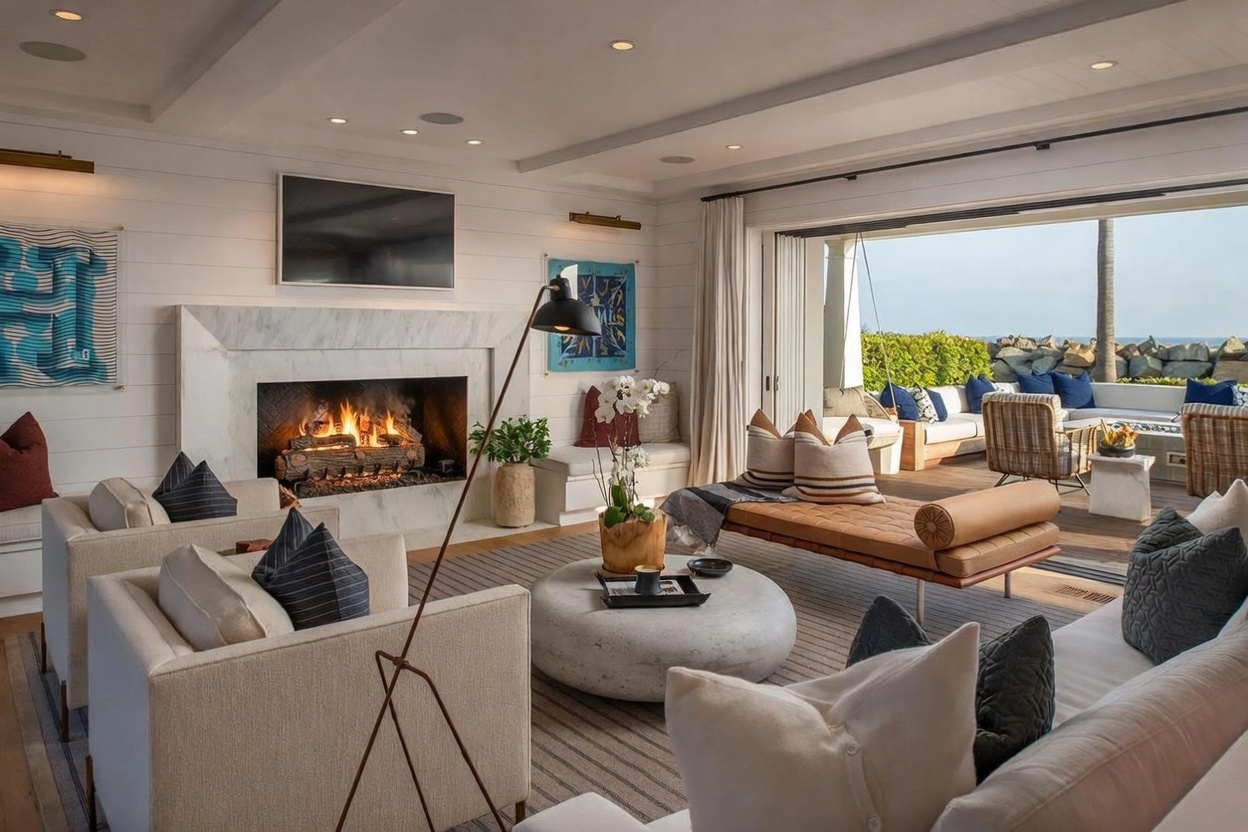 This bright and airy living room features a prominent stone-clad fireplace surround and a large, circular coffee table as a central focal point. The space is furnished with plush cream-colored seating and a leather daybed, seamlessly connecting to an expansive outdoor deck through wide-opening glass doors. The coastal-inspired design creates a sophisticated yet relaxed atmosphere, perfect for indoor-outdoor living with a view of the ocean.