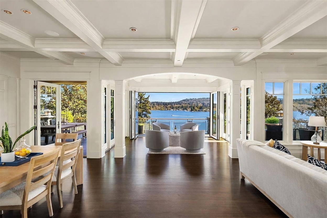 This elegant living area features a seamless transition to an outdoor deck through expansive folding glass doors that frame a stunning lake view. The room is defined by sophisticated coffered ceilings, rich dark hardwood flooring, and a neutral color palette that emphasizes the bright, airy atmosphere. A cozy seating arrangement with four plush armchairs creates an inviting focal point, while the adjacent dining space adds to the home's open-concept, luxury appeal.