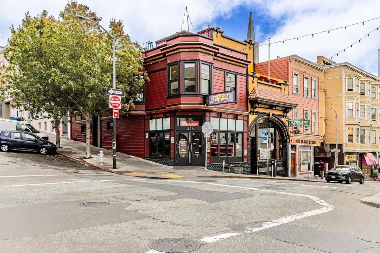 This image captures a prominent corner building in an urban setting, featuring a vibrant red facade with dark trim and a distinctive bay window on the upper level. The ground floor houses a commercial establishment, identified as 'Showdown,' with large glass windows and a central entrance. The perspective is taken from the street level, showcasing the building's architectural character alongside neighboring structures and a mature tree on the adjacent sloped street.