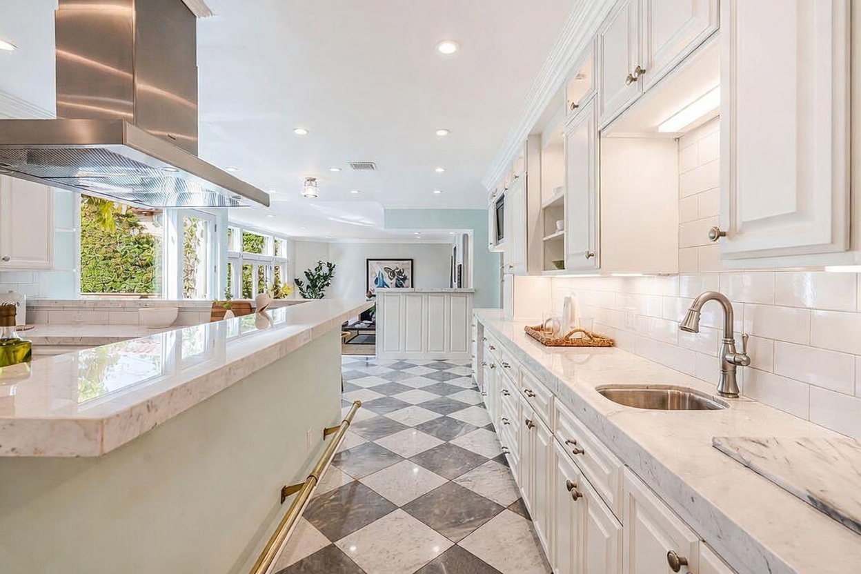 This bright, elegant kitchen features a long breakfast bar with a stone countertop and a brass foot rail, overlooking a spacious living area. The space is defined by white cabinetry, a subway tile backsplash, and a striking black-and-white checkered stone floor. A large stainless steel range hood serves as a focal point above the island, while recessed lighting and under-cabinet lighting create a warm, inviting atmosphere.