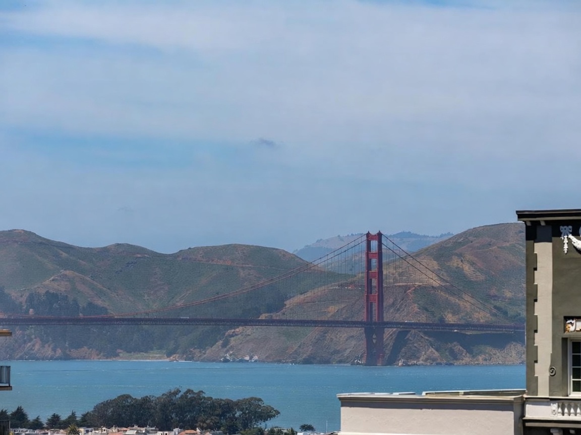 This image captures a stunning, elevated view of the Golden Gate Bridge from what appears to be a private balcony or terrace. The perspective highlights the iconic red structure set against the rolling hills of the Marin Headlands and the blue waters of the bay. To the right, a portion of the building's exterior wall is visible, featuring a classic architectural style with a window, suggesting a sophisticated residential setting.