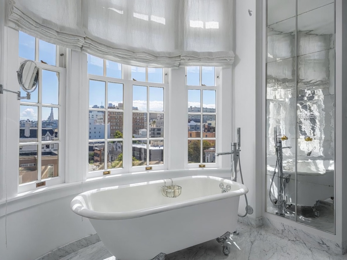 This elegant primary bathroom features a classic clawfoot soaking tub positioned in front of a large bay window that offers expansive city views. The space is finished with light-toned stone flooring and a mirrored wall that enhances the bright, airy atmosphere. A wall-mounted magnifying mirror and polished metal fixtures add a touch of refined detail to this sophisticated retreat.