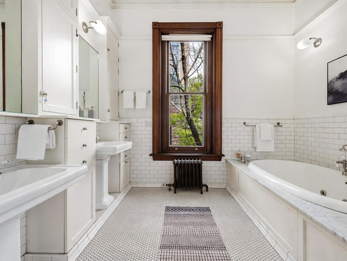 This elegant, classic-style bathroom features a double vanity with white cabinetry and a pedestal sink, complemented by white subway tile wainscoting and a patterned mosaic tile floor. A deep soaking tub is positioned under a window with a rich, dark wood frame, while a vintage-style radiator adds character to the space. The room is bright and airy, with wall-mounted lighting fixtures providing a warm, inviting atmosphere.