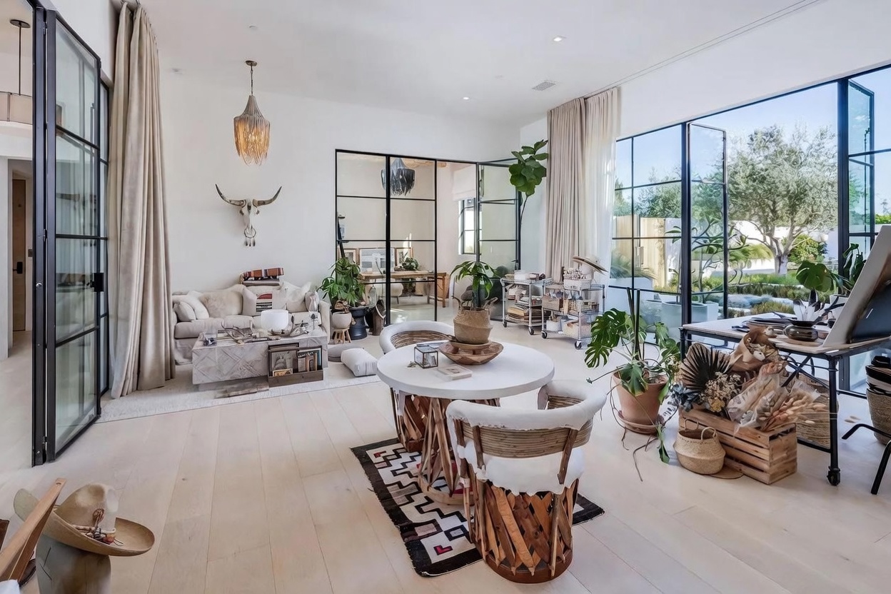 This bright, open-concept living and dining area features light wood flooring and a bohemian-inspired aesthetic with natural textures and indoor greenery. The space is anchored by a central round dining table with unique woven chairs, while a cozy seating area with a light-toned sofa and a decorative animal skull wall mount sits to the left. Large, black-framed glass doors and partitions provide a seamless transition to an adjacent room and the outdoor garden, creating an airy, light-filled atmosphere.