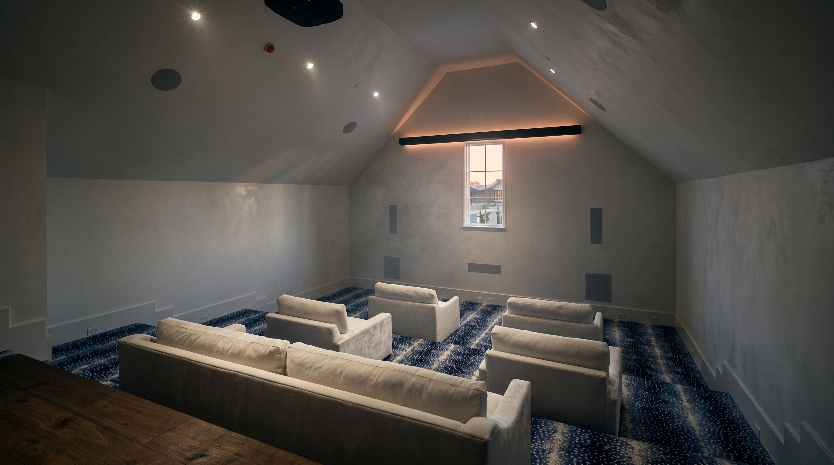 This dedicated home theater room is situated in an attic space, featuring a vaulted ceiling and a tiered floor layout for optimal viewing. The room is furnished with a plush sofa and four individual armchairs, all upholstered in light-colored fabric, facing a central window flanked by integrated wall speakers. A bold, blue-and-white patterned carpet adds a striking visual element to the minimalist, white-walled space, which is equipped with a ceiling-mounted projector.