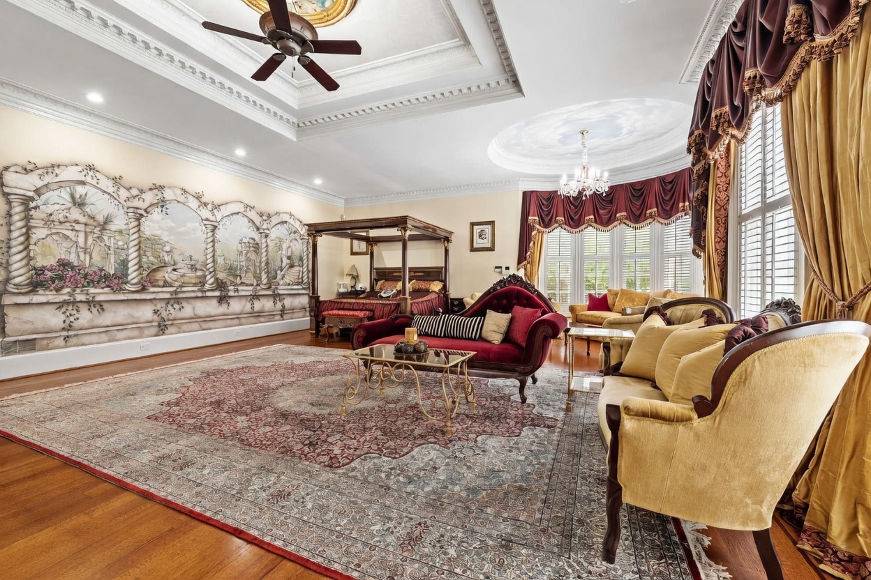 This expansive primary bedroom features a grand four-poster bed and a sophisticated seating area arranged on a large, ornate area rug. The room is distinguished by a detailed mural depicting classical architectural arches on the main wall, complemented by a coffered ceiling with a central fan and a separate circular tray ceiling area with a chandelier. Large windows draped in heavy, elegant curtains provide ample natural light, creating a luxurious and opulent atmosphere.