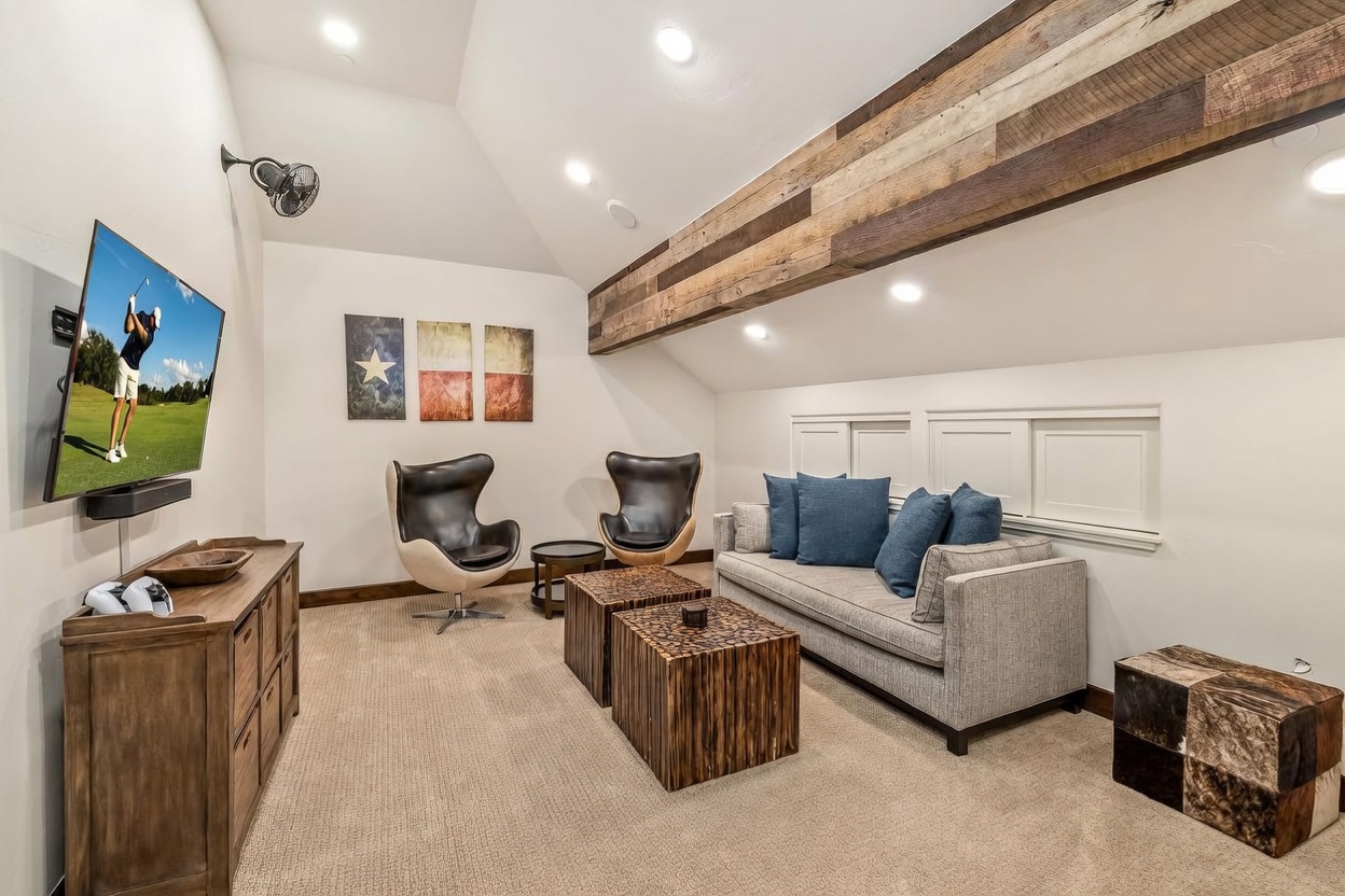 This cozy, attic-style living room features a vaulted ceiling accented by a prominent reclaimed wood beam. The space is furnished with a grey sofa, two modern leather swivel chairs, and two unique wooden block coffee tables, all set against a neutral backdrop with Texas-themed wall art. A large television is mounted on the wall, creating a comfortable media area under the sloped roofline.