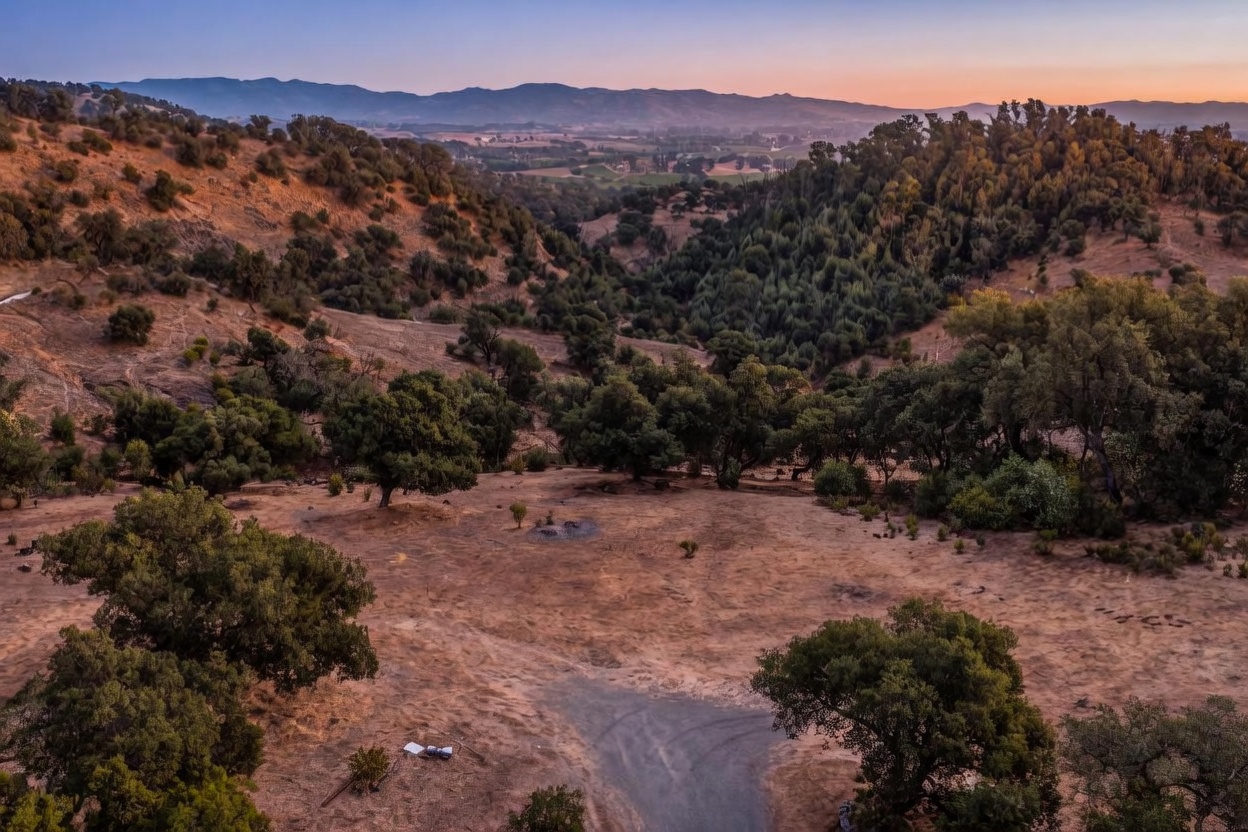 This elevated aerial view showcases a vast, sun-drenched landscape featuring rolling hills covered in golden grasses and scattered oak trees. The scene offers an expansive perspective of a rural or semi-rural property with gentle slopes and a distant valley, emphasizing the natural beauty and sense of privacy. It captures the serene, rustic character of the terrain, making it ideal for buyers looking for scenic acreage.