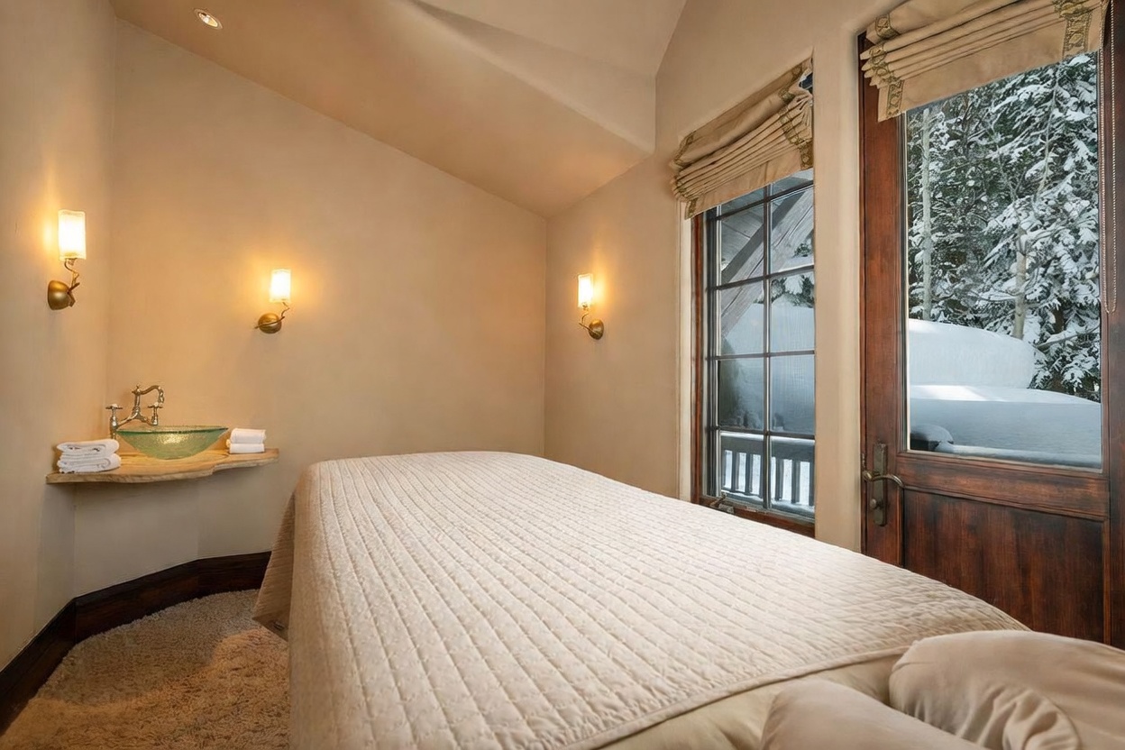 This serene spa or massage room features a plush treatment table centered in the space, illuminated by warm wall-mounted sconces. A unique glass vessel sink sits atop a natural-edge stone shelf, adding a rustic yet refined touch to the neutral-toned room. A large window with a Roman shade provides a picturesque view of a snow-covered forest, creating a tranquil and inviting atmosphere.