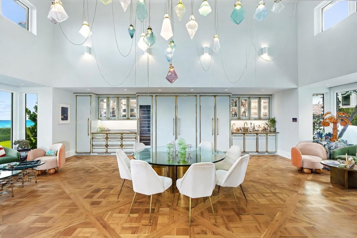 This elegant dining area features a central glass-topped table surrounded by white upholstered chairs, set against a backdrop of custom light-blue cabinetry with gold hardware. A striking installation of numerous geometric glass pendant lights hangs from the high ceiling, creating a dramatic focal point above the room. The space is finished with intricate patterned wood flooring and is flanked by comfortable seating areas, offering a sophisticated and airy atmosphere.