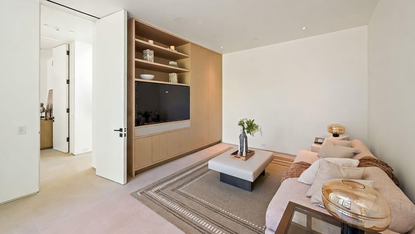 This modern living room features a sleek, light wood built-in media center with integrated shelving and a flat-screen television. A plush, neutral-toned sofa is adorned with decorative pillows, complemented by a low-profile square ottoman coffee table centered on a textured area rug. The minimalist aesthetic is enhanced by clean white walls and recessed lighting, creating a bright and inviting atmosphere.