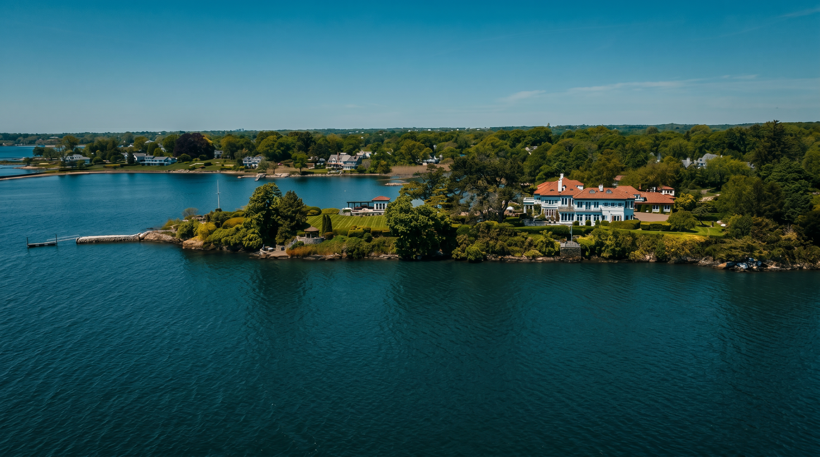 This expansive aerial view captures a grand waterfront estate situated on a lush, private peninsula surrounded by deep blue water. The property features a large, multi-story residence with a classic architectural style, a sprawling manicured lawn, and a private dock extending into the bay. The surrounding landscape is densely wooded, emphasizing the secluded and prestigious nature of this coastal retreat.