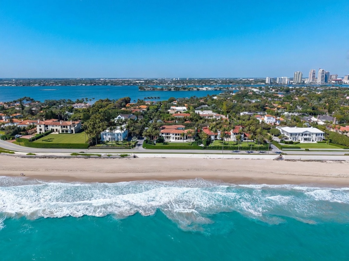 This high-altitude aerial perspective captures a prestigious coastal estate situated between the turquoise Atlantic Ocean and a calm inland waterway. The property features a sprawling Mediterranean-style residence with a terracotta roof, surrounded by lush tropical landscaping, manicured lawns, and mature palm trees. The composition highlights the prime waterfront location, showcasing the expansive sandy beach in the foreground and the distant city skyline across the bay.
