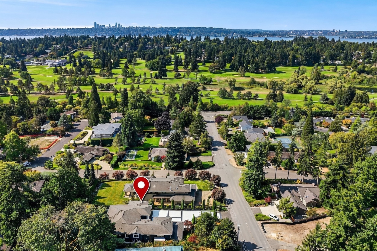 This high-angle aerial view captures a sprawling residential property situated adjacent to a lush, expansive golf course. The home features a complex, multi-gabled roofline and a well-manicured lawn, with a red location pin highlighting the residence's position within the neighborhood. In the distance, a scenic lake and a city skyline are visible under a clear blue sky, emphasizing the property's desirable location and open surroundings.