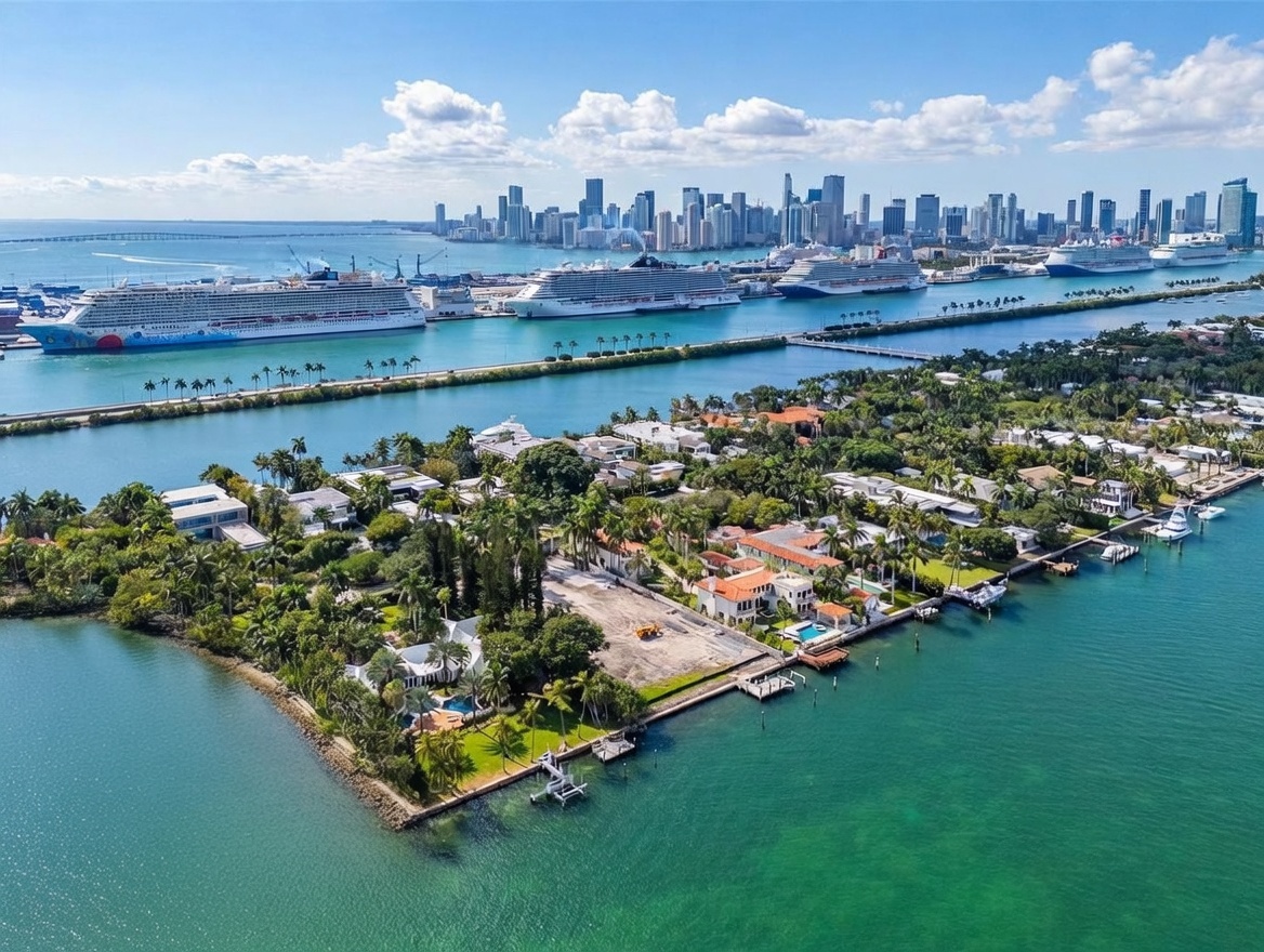 This high-altitude aerial view captures a luxurious waterfront residential neighborhood on a tropical island, characterized by expansive estates with private docks and swimming pools. In the background, the vibrant Miami skyline rises above the horizon, while several large cruise ships are docked in the deep blue channel separating the residential area from the city. The perspective provides a grand, cinematic overview of the prime coastal location, emphasizing the proximity to both maritime activity and urban amenities.