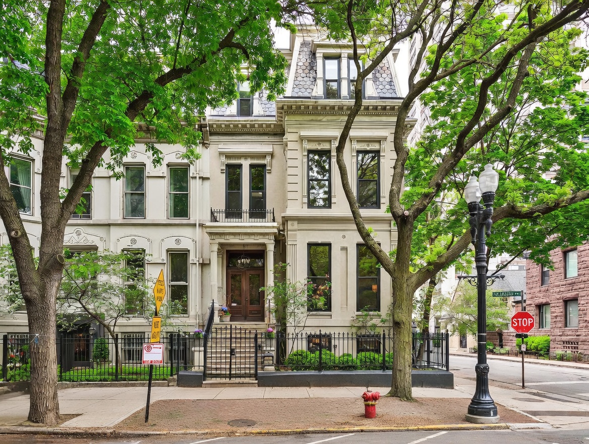 This elegant, multi-story historic townhouse features a light-colored stone facade with a prominent mansard roof and detailed architectural molding. The front entrance is elevated with a short staircase, framed by a black iron fence that encloses a small front garden. Mature trees frame the property, providing a lush, shaded streetscape in a classic urban neighborhood setting.