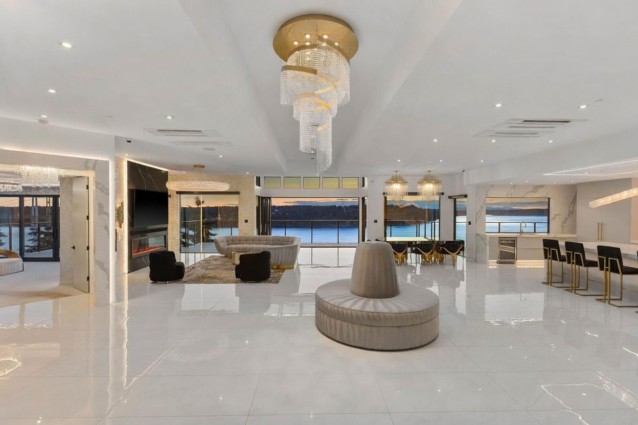 This expansive, open-concept living area features polished white tile flooring and a dramatic, multi-tiered crystal chandelier as a central focal point. The space includes a curved sofa arrangement, a unique circular lounge piece in the foreground, and a sleek kitchen area with bar seating to the right. Large glass doors provide an unobstructed view of the water, creating a seamless transition between the luxurious interior and the natural landscape outside.