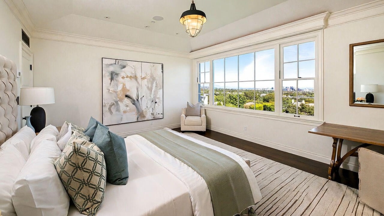 This spacious primary bedroom features a neutral color palette, a large bed with patterned accent pillows, and a soft area rug over dark wood flooring. A large window wall provides abundant natural light and expansive views of the surrounding landscape and city skyline. The room is accented by a large abstract painting, a cozy armchair in the corner, and a simple wooden desk, creating a serene and sophisticated atmosphere.