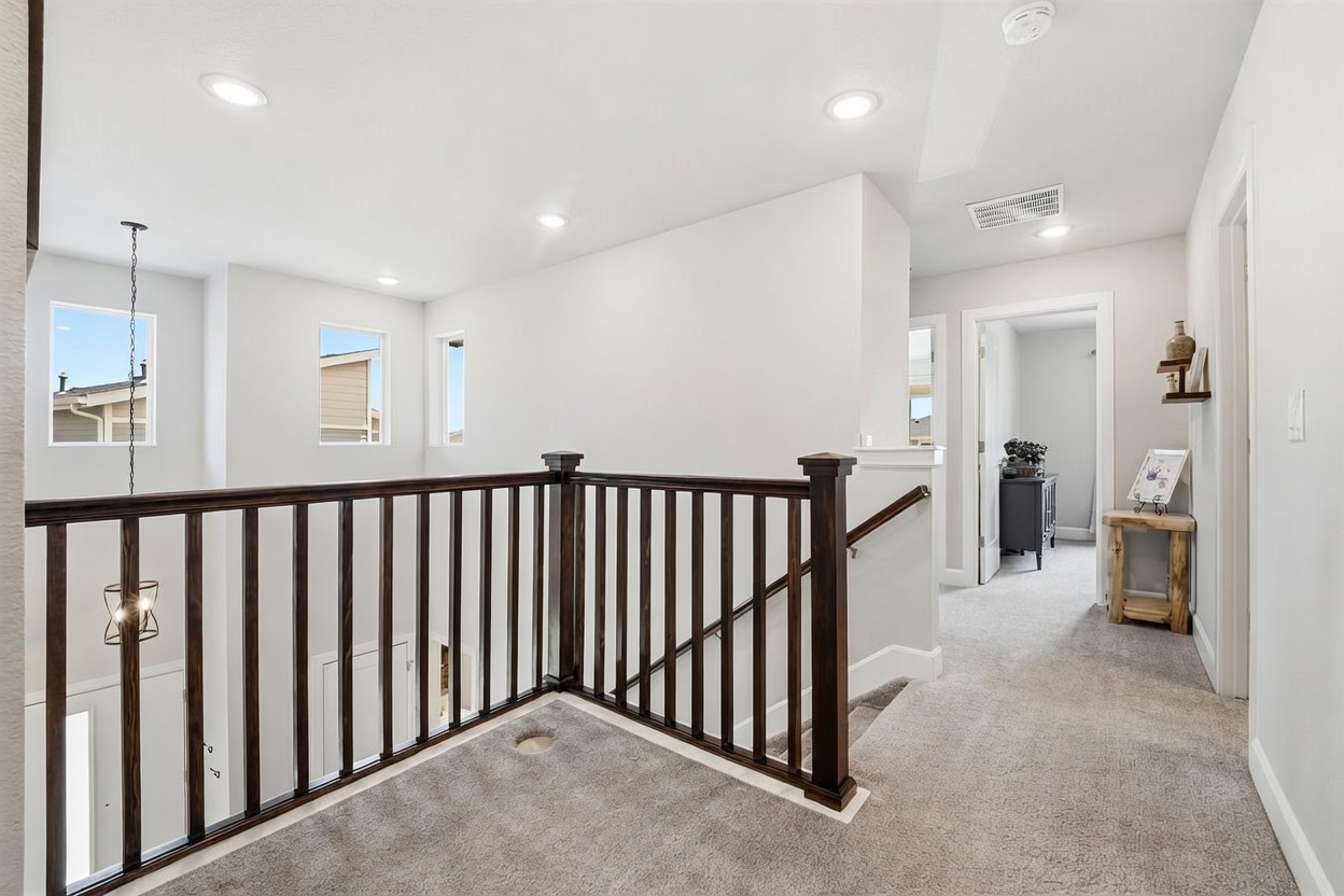 This image captures a bright, carpeted landing area at the top of a staircase, featuring a dark wood railing that overlooks the lower level. The space is illuminated by recessed lighting and natural light streaming through several windows, creating an open and airy atmosphere. A hallway leads toward the back of the home, where a small wooden console table with a framed picture adds a touch of decor to the neutral-toned interior.
