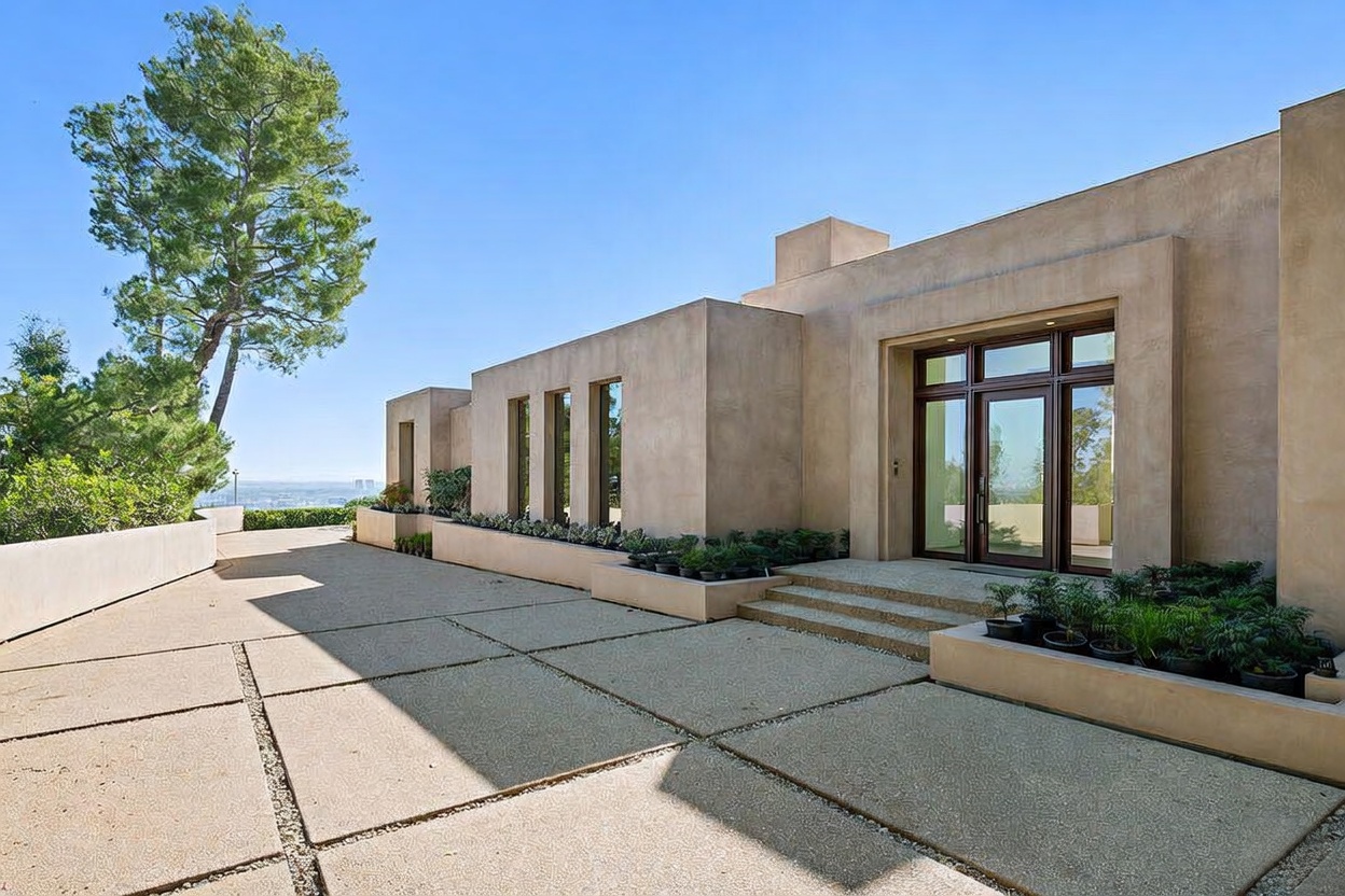 This striking modern entryway features a minimalist, earth-toned stucco facade with clean geometric lines and a prominent glass-paneled front door. A wide, textured concrete driveway leads up to the entrance, flanked by low-profile planters filled with lush greenery. The perspective captures the architectural symmetry and the expansive, open feel of the property, highlighting its sophisticated contemporary design.