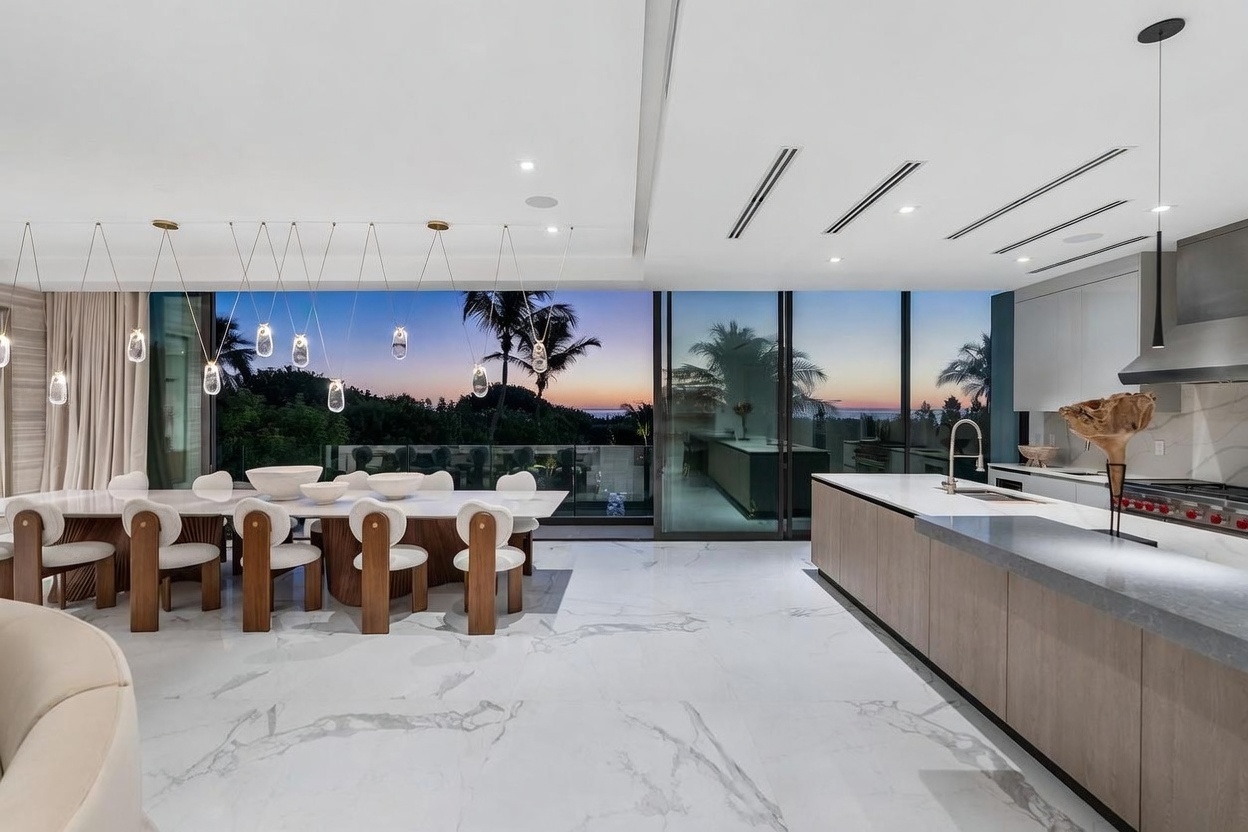 This open-concept space seamlessly blends a modern kitchen with a formal dining area, characterized by sleek cabinetry and a large island with a stone countertop. The room features expansive floor-to-ceiling glass walls that provide a panoramic view of the sunset over the trees and ocean beyond. A series of unique, sculptural pendant lights hangs above the dining table, creating a sophisticated and airy atmosphere.