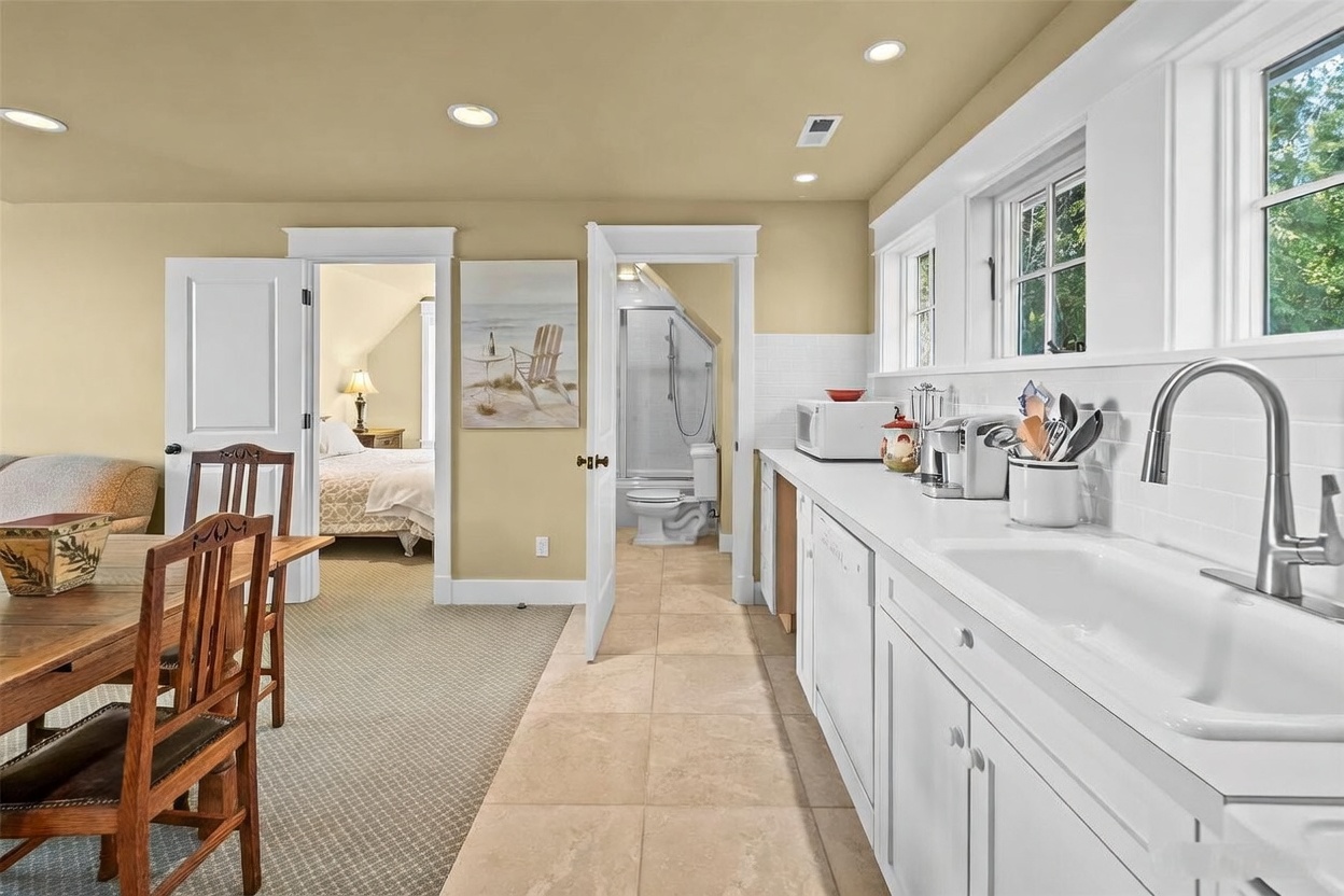 This interior view captures a functional kitchen space that transitions into a dining area and a bedroom beyond. The kitchen features white cabinetry, a white farmhouse-style sink, and a tiled backsplash, while the adjacent dining area is furnished with a wooden table and chairs. The perspective looks through an open doorway into a bedroom with a bed and a glimpse of an attached bathroom, creating an open and connected floor plan.