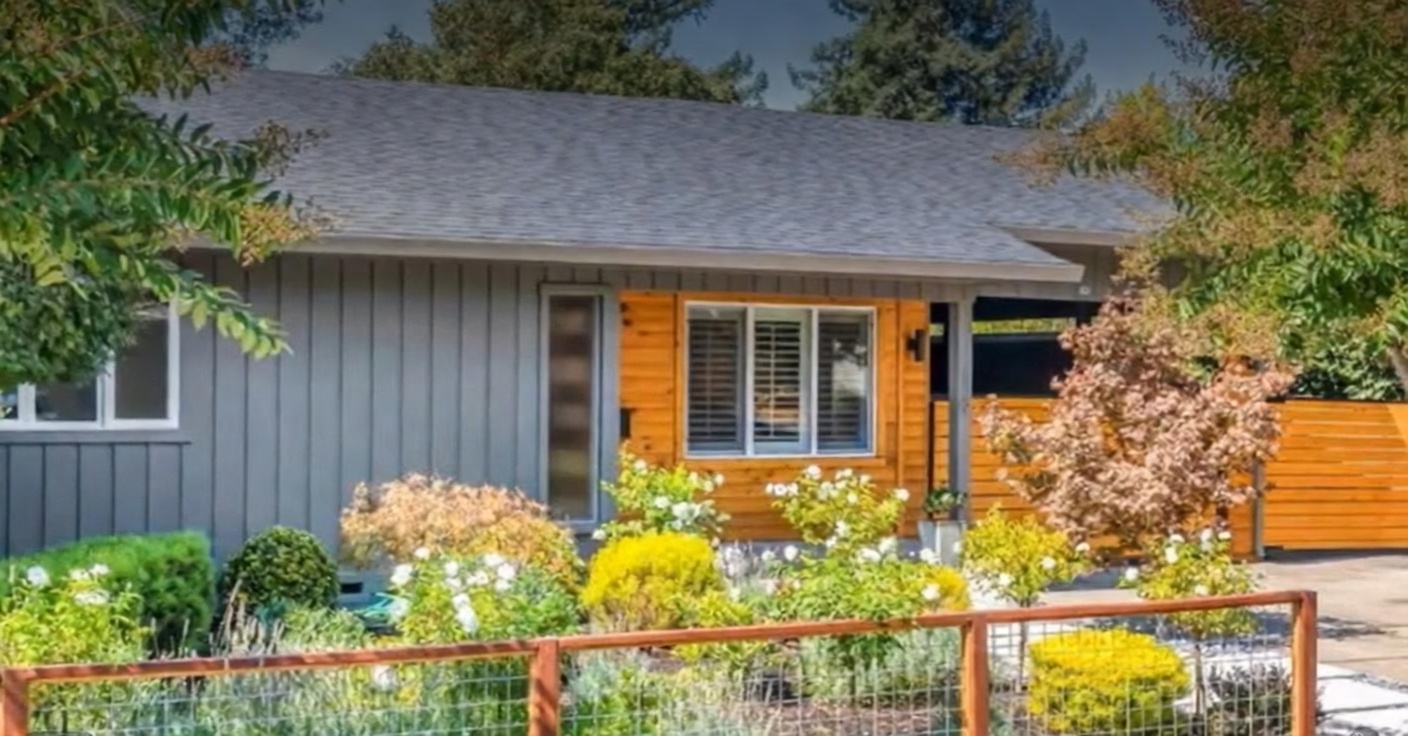 This charming single-story home features a modern mix of grey vertical siding and warm, natural wood paneling. The property is well-landscaped with a variety of shrubs and flowering plants, enclosed by a contemporary wire fencing that adds to the home's inviting curb appeal. A wide roofline and clean lines highlight the house's updated mid-century aesthetic.