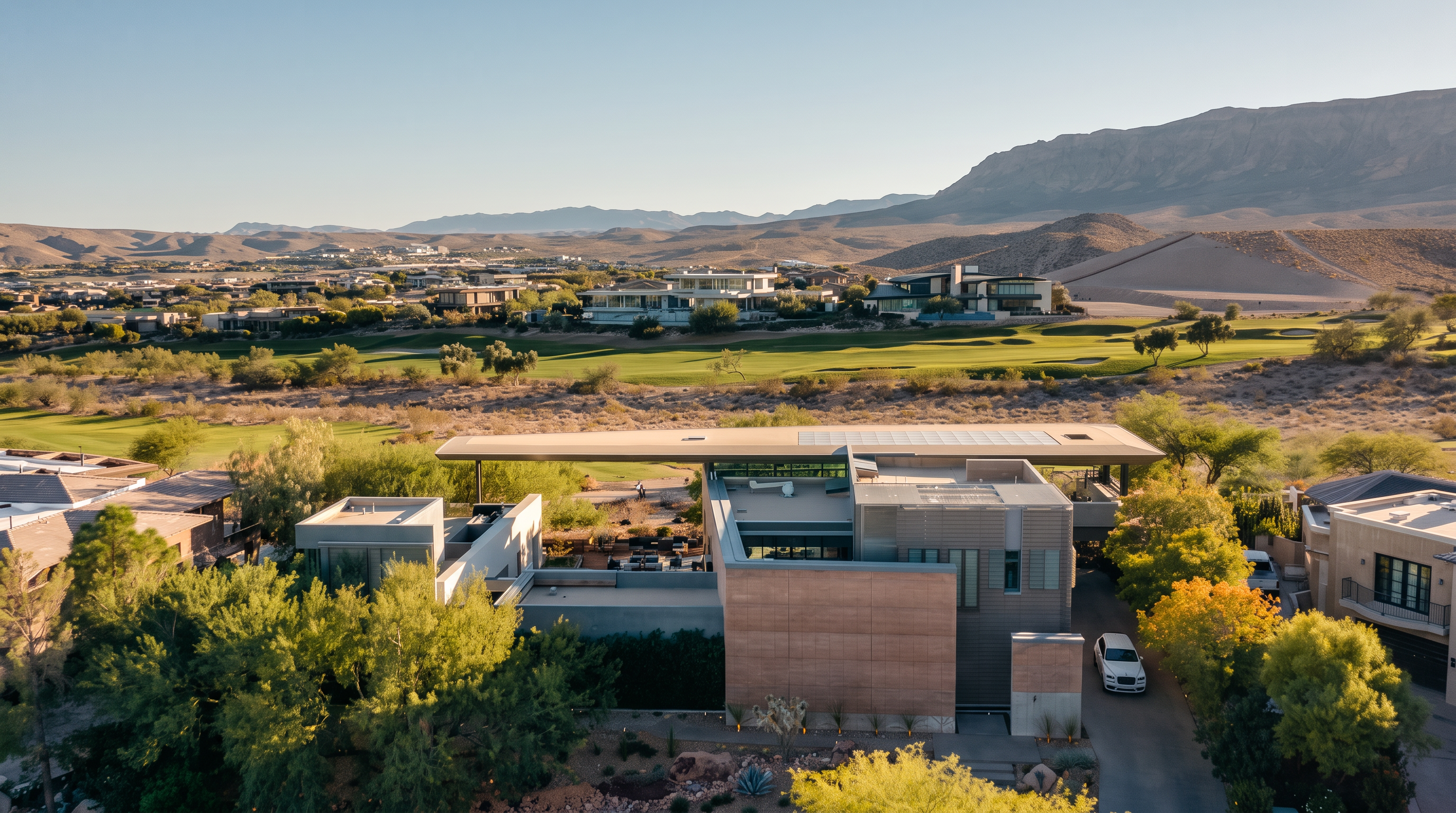 This high-angle aerial view captures a modern, multi-level residence set against a backdrop of desert mountains and a sprawling golf course. The home features a flat, expansive roofline, clean geometric lines, and a mix of stone and glass elements, with a private courtyard area visible in the center. The surrounding landscape is characterized by manicured green fairways and arid, rugged terrain, emphasizing the property's secluded and scenic location.