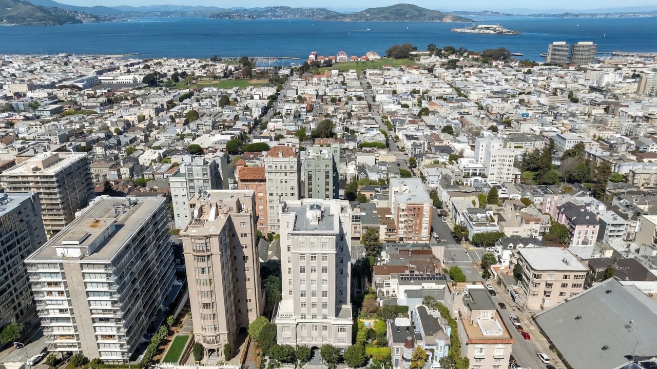 This high-angle aerial view captures a dense urban neighborhood in San Francisco, showcasing a mix of historic and modern high-rise residential buildings. The perspective looks out toward the San Francisco Bay, with Alcatraz Island visible in the distance under a clear blue sky. The scene highlights the city's characteristic grid layout, varied architectural styles, and proximity to the waterfront.