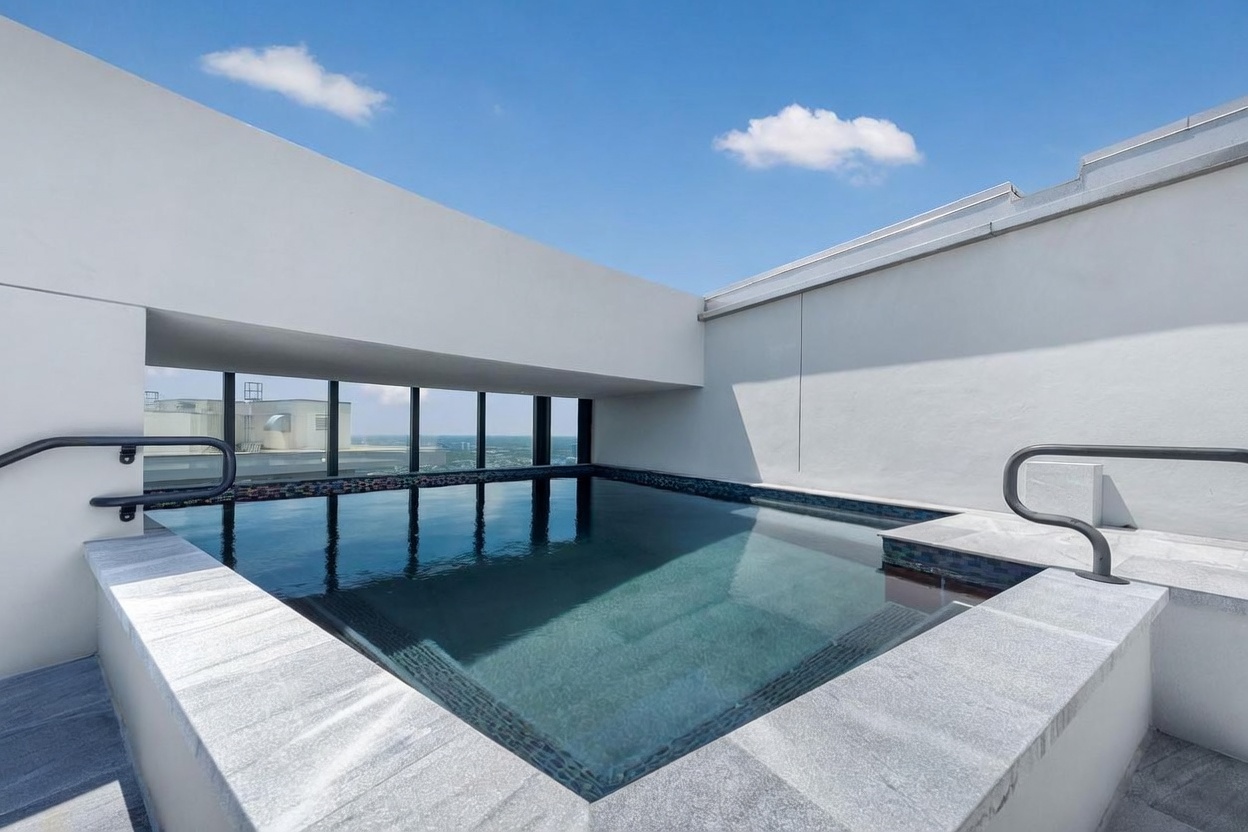 This elevated rooftop pool features a sleek, modern design with dark-toned tiling and a stone-surround edge. The pool is framed by a minimalist white architectural overhang and large windows that offer an expansive view of the surrounding area. The scene is bathed in bright daylight, creating a serene and luxurious atmosphere.