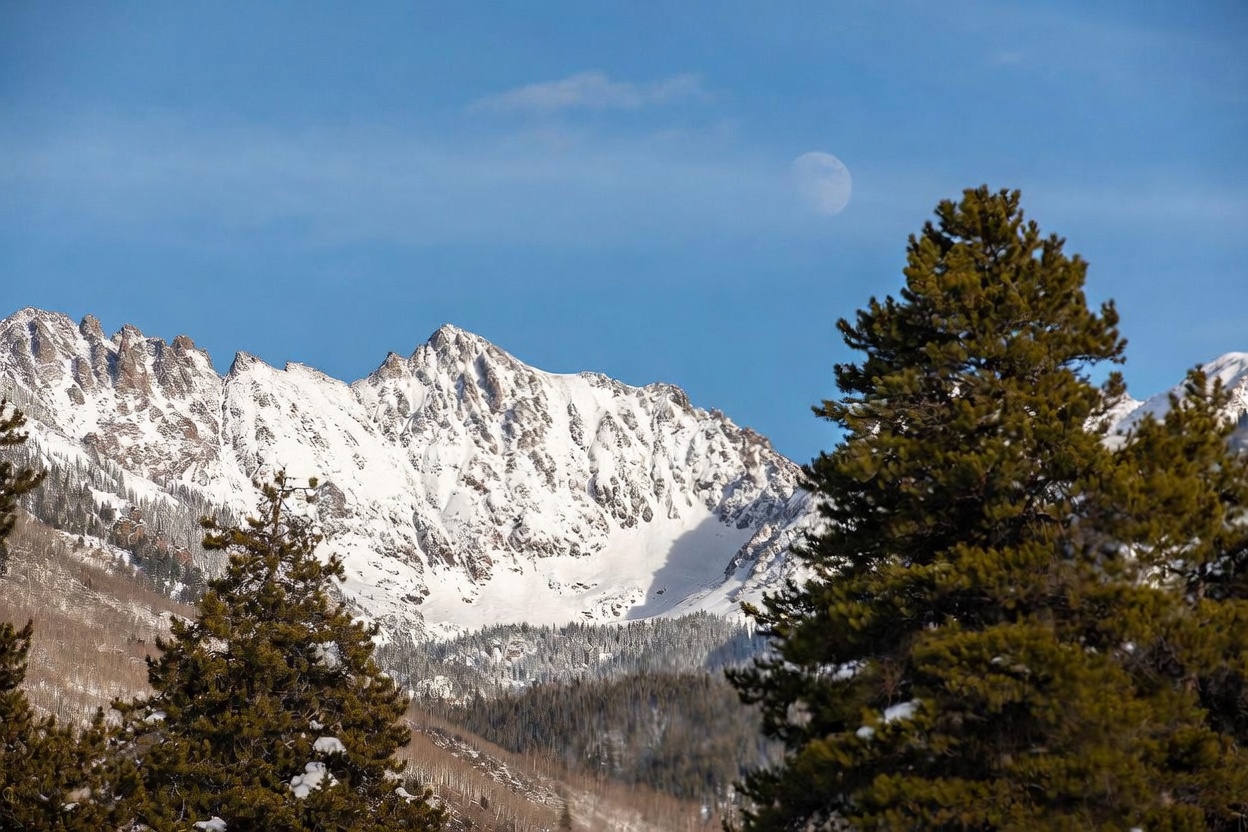 This scenic landscape shot captures a majestic, snow-capped mountain range under a clear blue sky, with a faint moon visible in the upper center. In the foreground, large, mature evergreen trees frame the view, adding depth and a sense of natural scale to the composition. The image evokes a serene, alpine atmosphere, highlighting the property's proximity to dramatic mountain vistas.