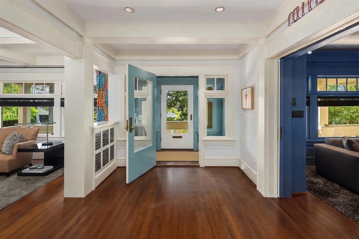 This bright and inviting entryway features rich, dark wood flooring and crisp white trim, creating a classic and welcoming atmosphere. A light blue door stands open to reveal a glimpse of the exterior, while the space connects seamlessly to adjacent living areas through wide, open doorways. The architectural details, including the paneled radiator cover and clean lines, suggest a well-maintained home with a blend of traditional and modern aesthetic elements.