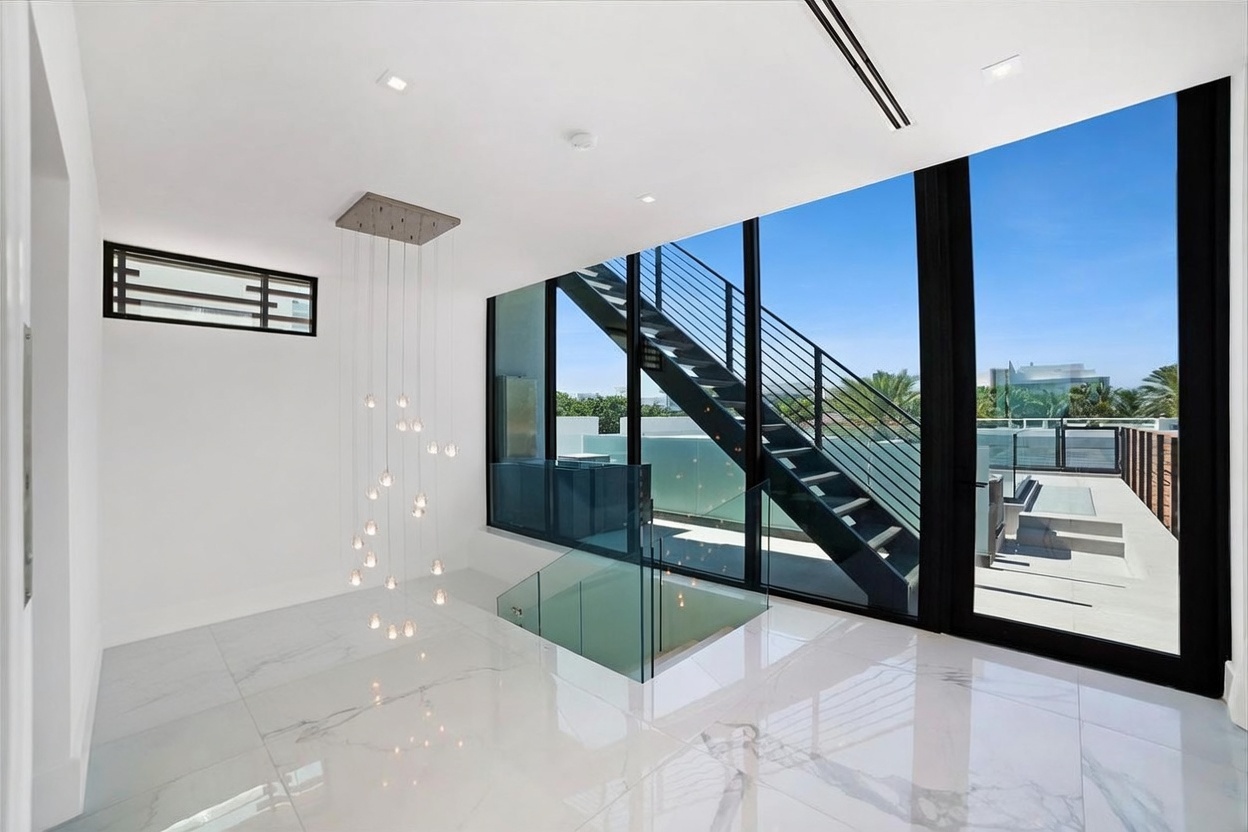 This modern, high-ceilinged interior space features a striking staircase with a glass railing and dark metal accents. A dramatic, multi-tiered pendant light fixture hangs in the stairwell, while large, floor-to-ceiling windows provide a seamless transition to an outdoor terrace. The polished, light-colored flooring reflects the abundant natural light, creating an airy and sophisticated atmosphere.