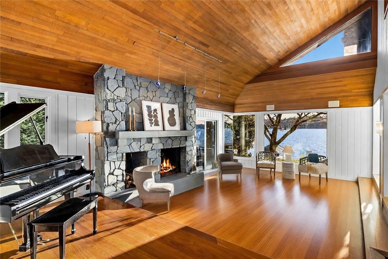 This spacious, light-filled living room features a striking floor-to-ceiling stone fireplace and a dramatic vaulted wood-paneled ceiling. A grand piano sits prominently in the foreground, while large windows offer expansive, serene views of the lake and surrounding trees. The room is elegantly furnished with a comfortable sofa, a modern coffee table, and accent chairs, creating a sophisticated yet cozy atmosphere.