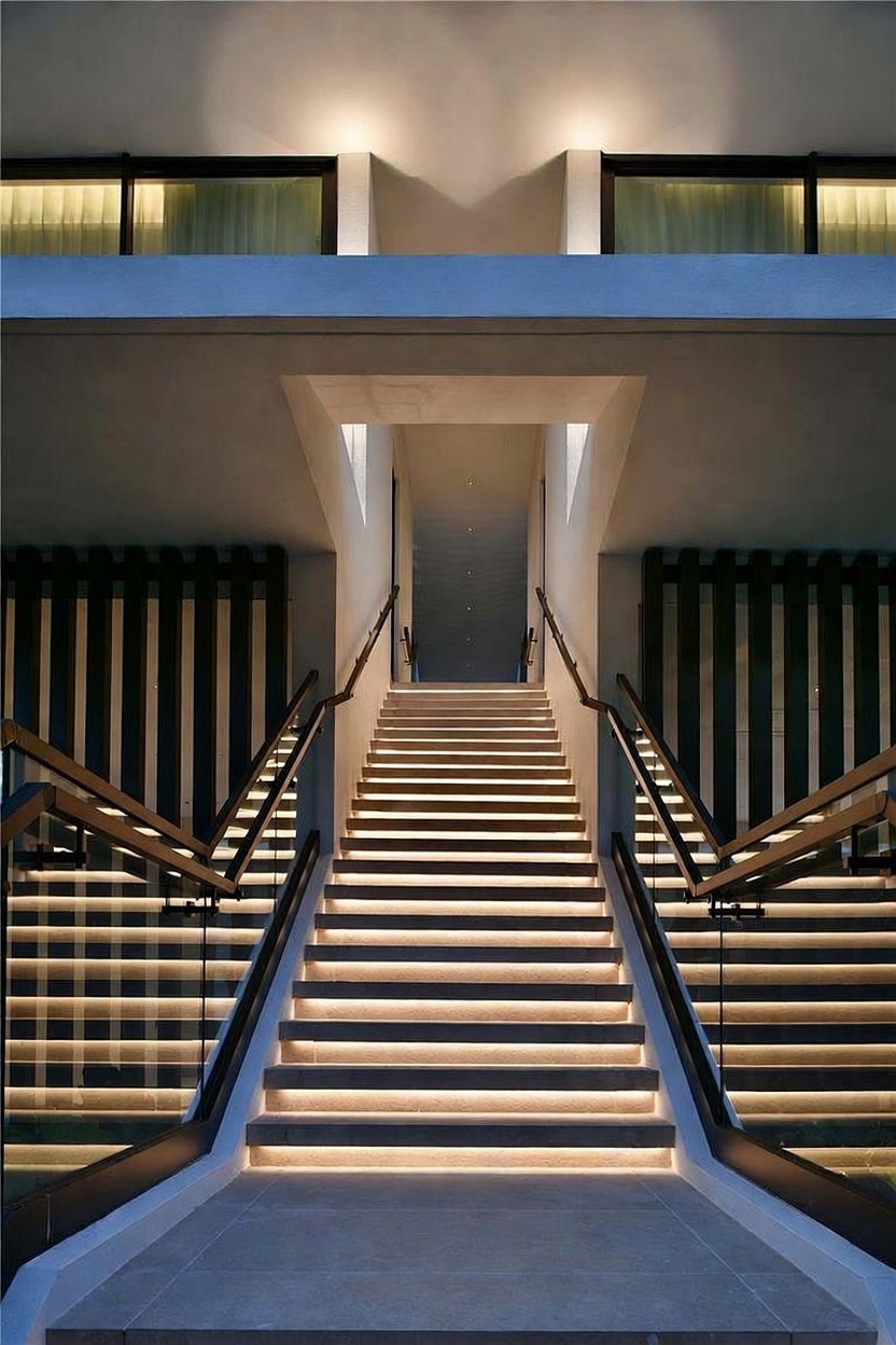 This striking staircase features a symmetrical design with illuminated steps that create a warm, inviting glow. The modern aesthetic is defined by clean lines, glass railings with dark metal handrails, and vertical slat accents on either side. The perspective is centered, drawing the eye upward toward a bright, minimalist landing that suggests a sophisticated and contemporary architectural style.