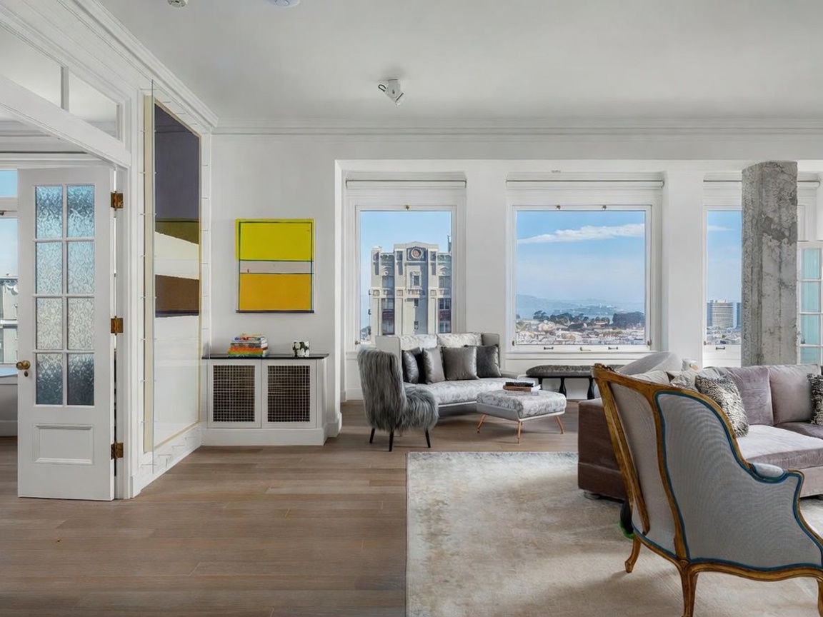 This bright and airy living room features light wood flooring and a sophisticated, eclectic design aesthetic. Large windows frame expansive city and bay views, while a mix of modern art and classic furniture pieces creates a refined atmosphere. The space is anchored by a plush area rug and includes a unique textured accent chair and a custom-built storage console.