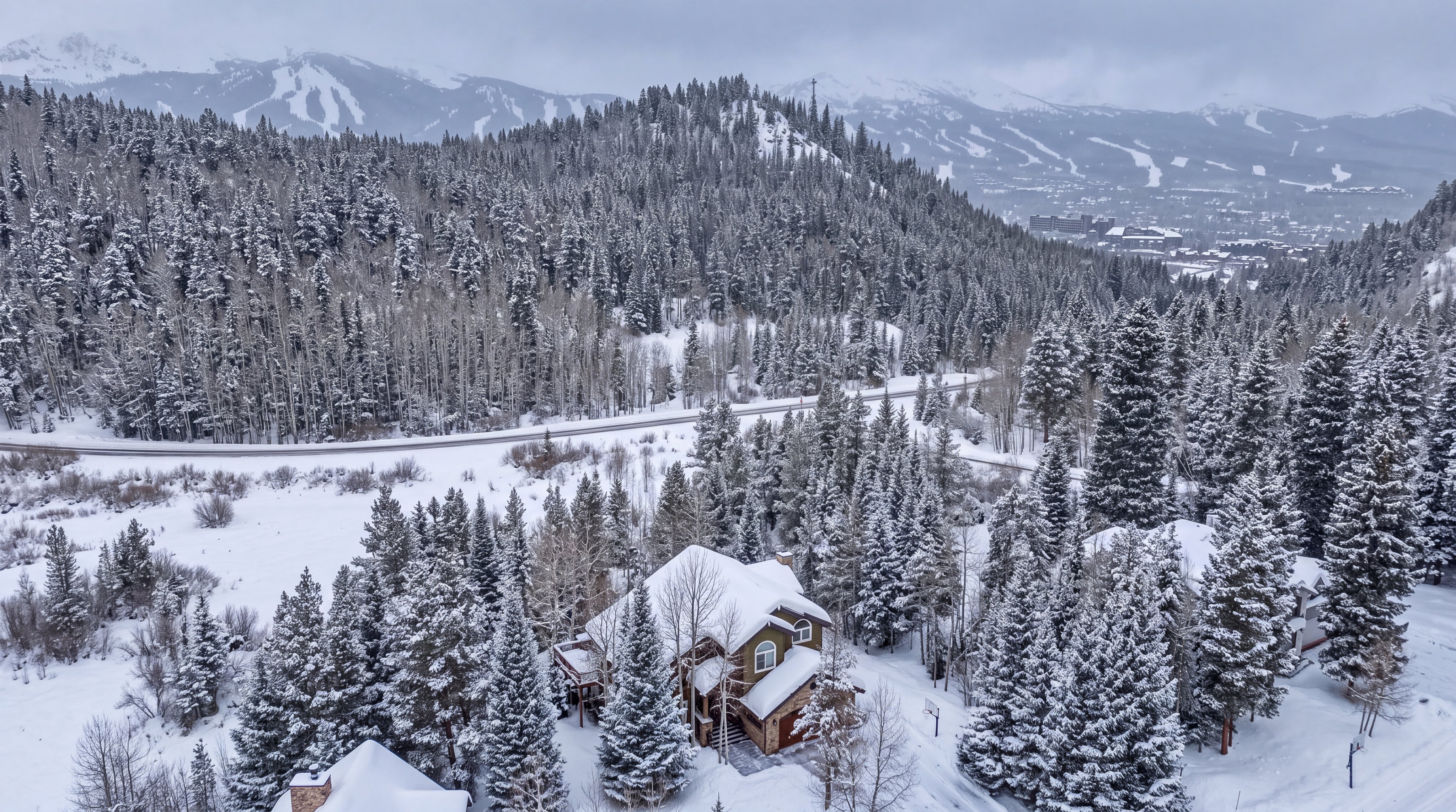 This aerial view captures a mountain home nestled in a snow-covered forest, showcasing its quintessential alpine setting. The property features traditional architectural details, including a steeply pitched roof designed to shed snow, and a large garage, all surrounded by a serene, wintry landscape. Its location near a main road and proximity to a ski resort make it an ideal retreat for winter sports enthusiasts.