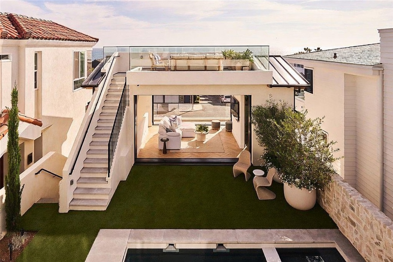 This rear view captures a modern coastal home featuring a seamless indoor-outdoor transition with large sliding glass doors opening to a living area. The property includes a manicured artificial turf lawn, a swimming pool in the foreground, and an external staircase leading to a spacious rooftop deck furnished with lounge seating. The architectural style is clean and contemporary, emphasizing light-colored finishes and a bright, airy atmosphere.